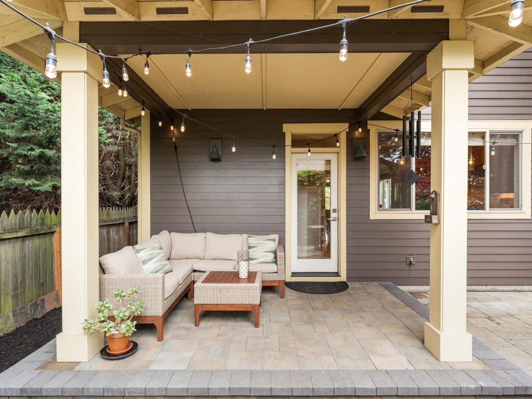 A cozy outdoor patio with a sectional sofa, coffee table, and string lights hanging from a wooden pergola. Ideal for Portland Oregon real estate enthusiasts, the space features a tiled floor and potted plant, with dark siding and a door to the house in the background.