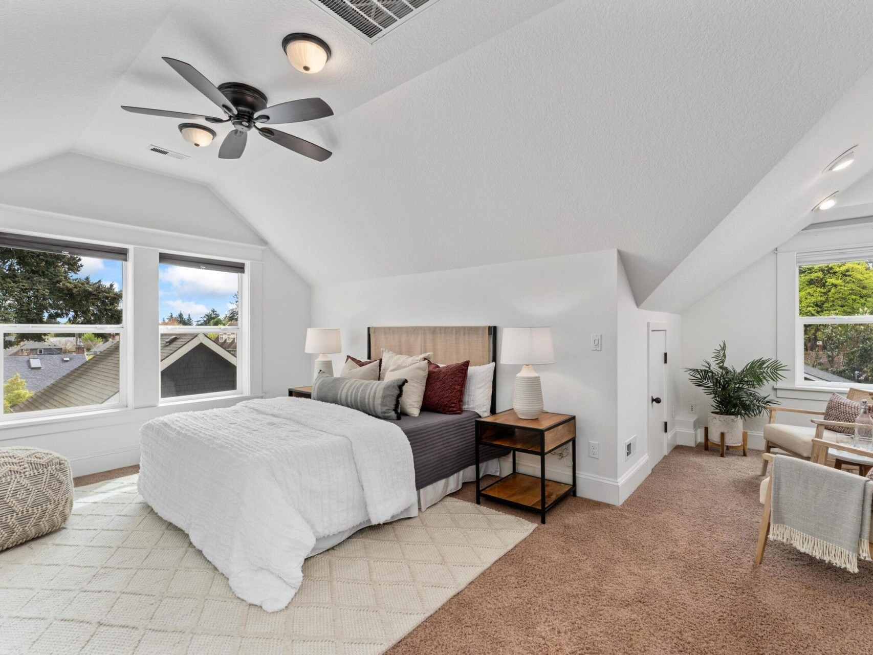 A cozy attic bedroom in Portland, Oregon real estate with a sloped ceiling features a bed with white and gray bedding, two bedside tables with lamps, a brown carpet, and a seating area with chairs. Large windows let in natural light, while a ceiling fan gently spins above.