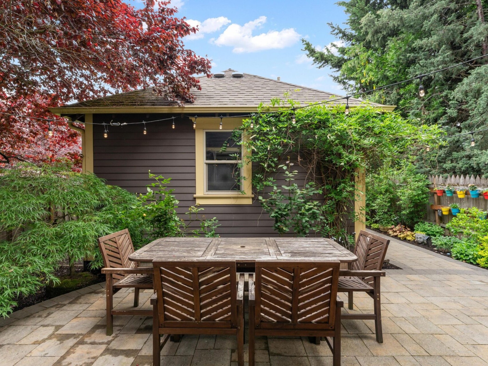 A cozy backyard in Portland, Oregon real estate features a wooden table and four chairs on a stone patio. String lights hang above, surrounded by lush greenery. A small brown shed with a window and yellow trim sits in the background, perfectly curated by your top Portland realtor.