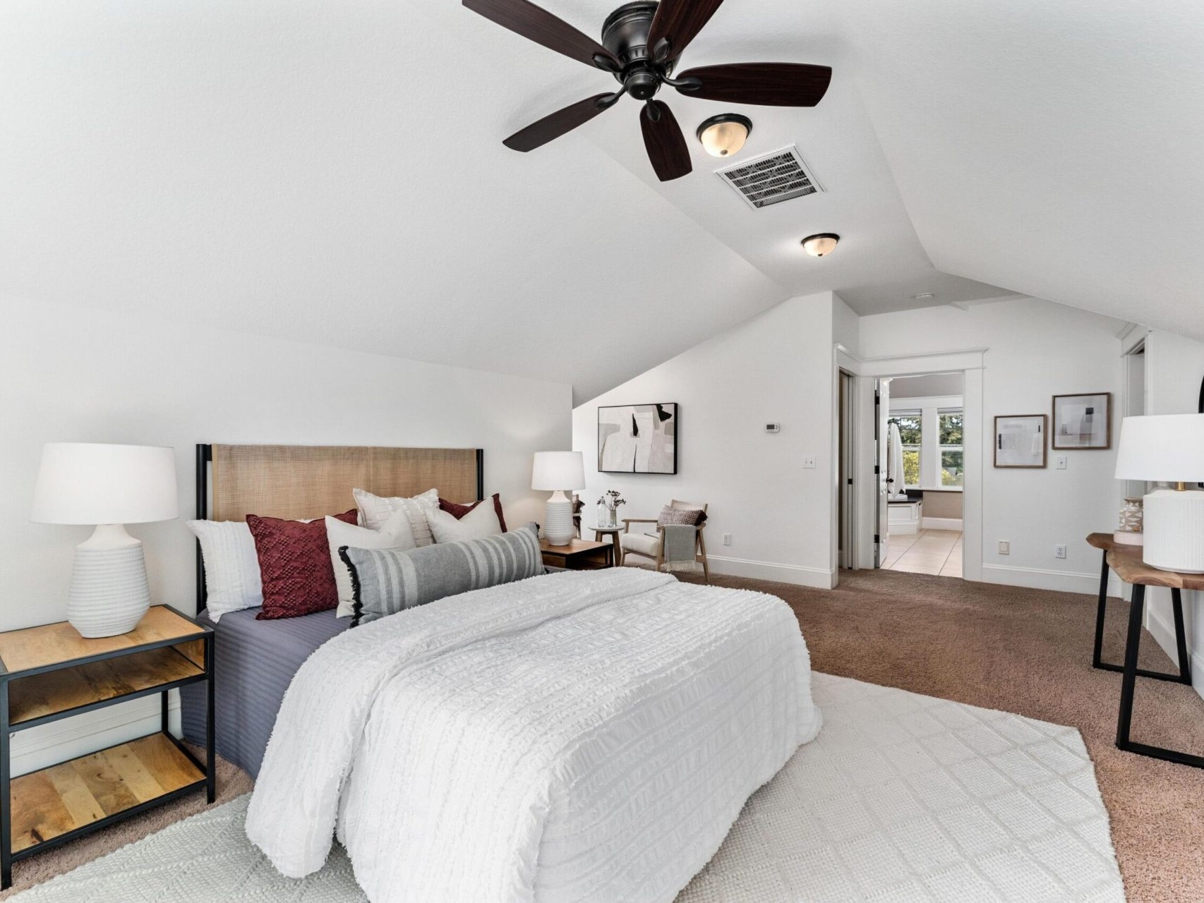 A cozy bedroom in a Portland Oregon real estate gem, featuring a large bed with white and dark red pillows, twin bedside tables with lamps, a ceiling fan, and a soft beige carpet. A console table with a decorative mirror and a view of the hallway complete the serene atmosphere.