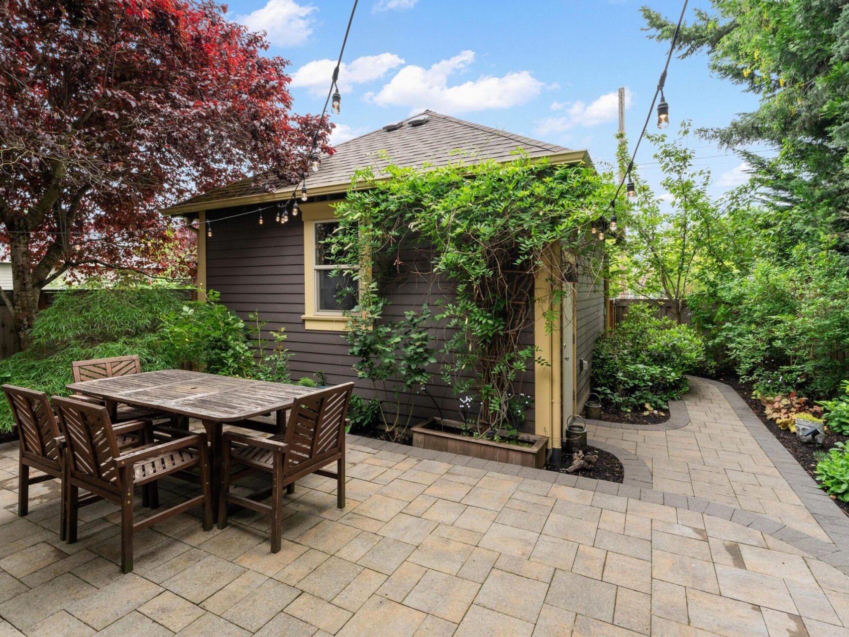 A cozy backyard in Portland, Oregon Real Estate features a small brown shed with green vines growing on it. There's a wooden table with four chairs on a paved patio. Surrounding the space are trees and shrubs, creating a lush garden atmosphere. String lights hang above the area.