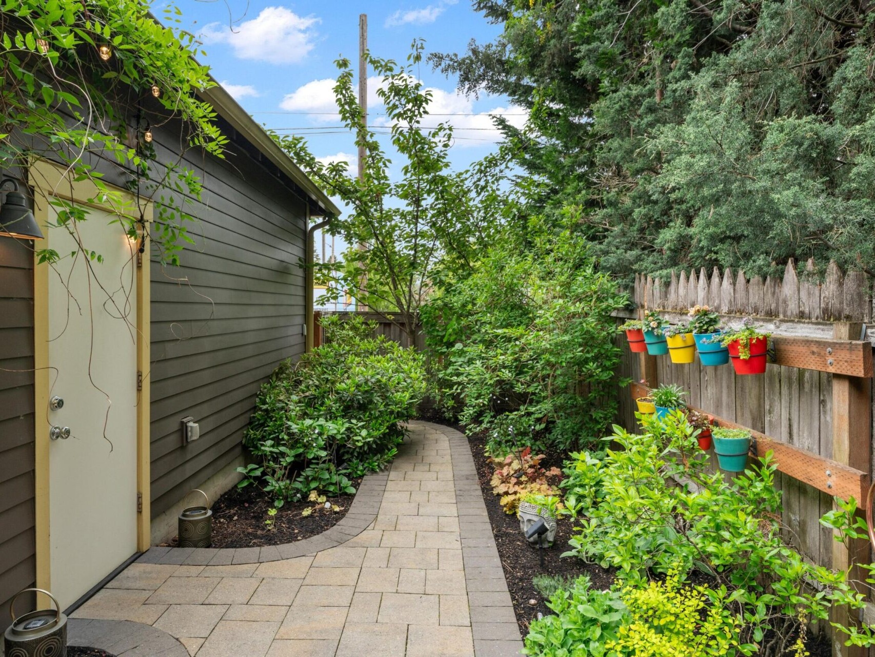 A narrow garden path lined with greenery and shrubs leads to a beige door, showcasing the charm of Portland Oregon real estate. Colorful potted plants hang on a wooden fence to the right, framed by tall bushes and a clear sky in the background.