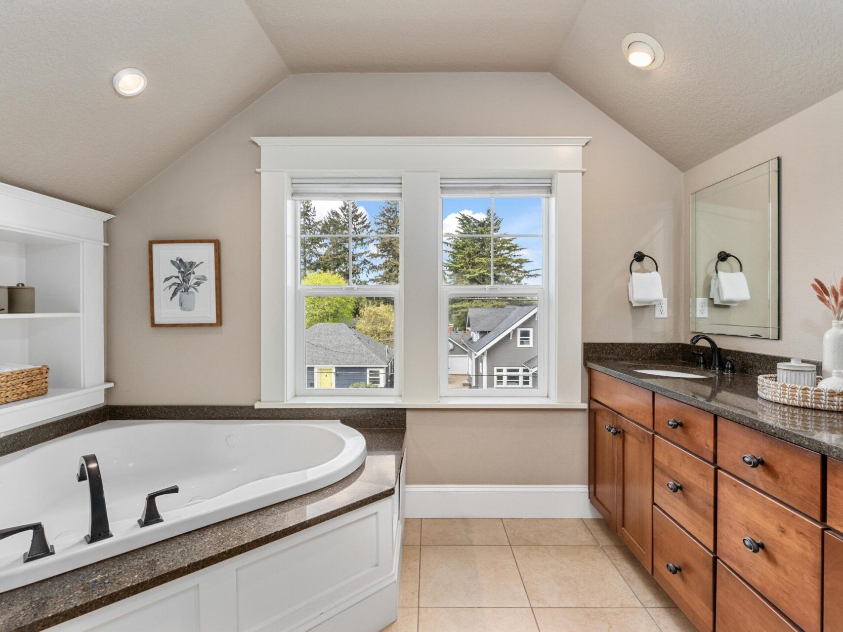 A bathroom in a Portland Oregon Real Estate listing features a large bathtub and brown wooden cabinets. There's a window with views of trees and neighboring houses. The countertop includes a sink, decorative items, and open shelves with baskets and a framed picture.