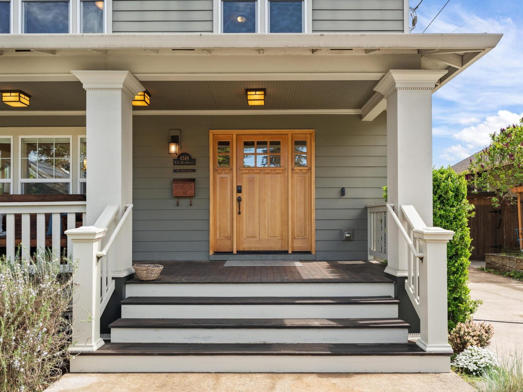 The front view of a house features a welcoming wooden door, wide porch, and white railings. Flanked by shrubs and trees, the inviting entrance is enhanced by ceiling lights and a mailbox. Explore Portland Oregon Real Estate for more bright and charming exteriors like this one.