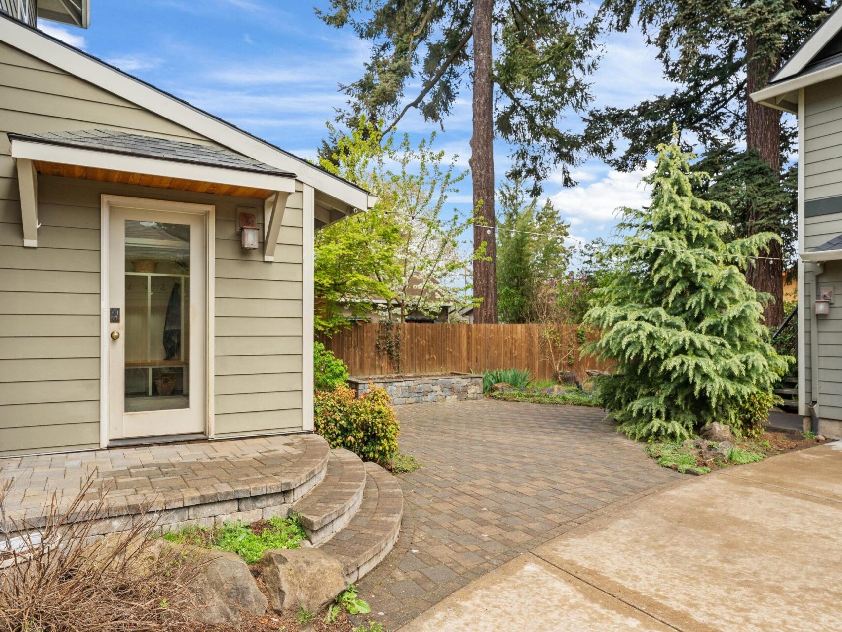 A small, paved courtyard nestled between two houses with beige siding, surrounded by lush greenery. The yard, common in Portland Oregon real estate, features a wooden fence and a mix of mature trees and shrubs. A clear blue sky stretches above, inviting you to relax in this serene space.