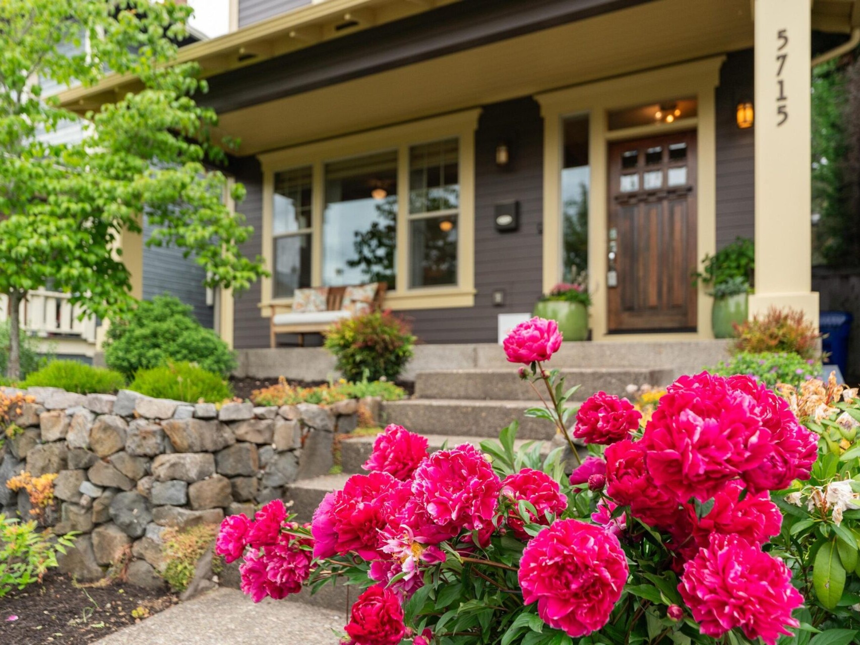 A charming house with a wooden front door and large windows is nestled within lush greenery. Bright pink peonies blossom vibrantly near a stone wall, adding color to the scene. For more homes like this, trust your top Portland Realtor to guide you through the Portland Oregon real estate market.