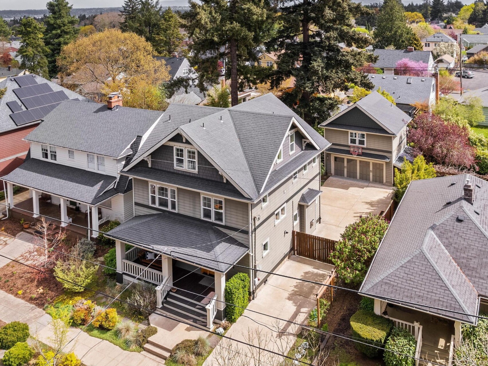 Aerial view of a Portland Oregon real estate neighborhood showcasing a large gray house with a gabled roof and front porch, surrounded by trees and landscaped gardens. Nearby, other homes boast solar panels on their roofs, a highlight often emphasized by top Portland Realtors.