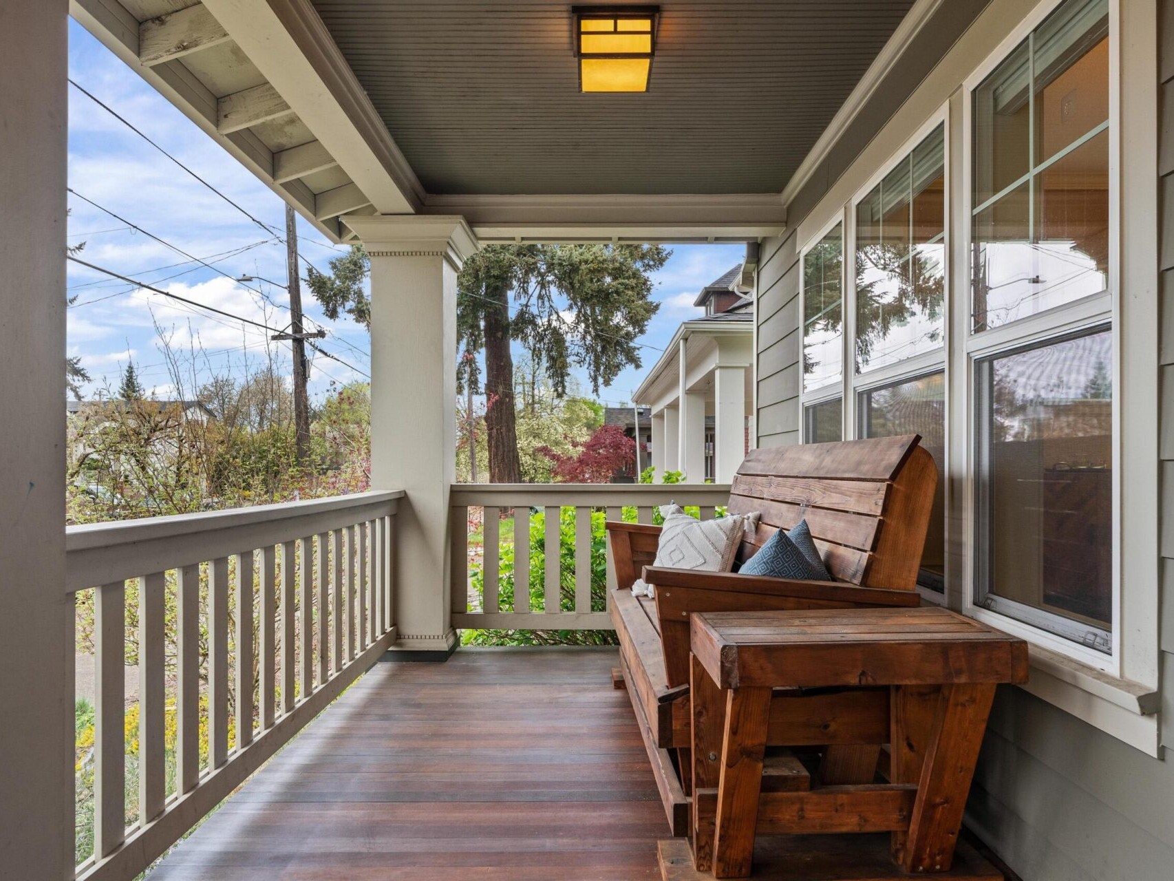 A cozy wooden porch with a rustic bench, pillows, and a covered roof offers a serene and inviting atmosphere. Overlooking trees, power lines, and neighboring houses, it's a charming slice of Portland Oregon real estate that captivates with its tranquil setting.