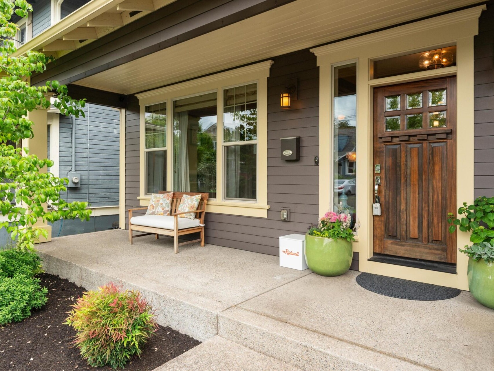 A welcoming front porch with gray siding and a wooden door invites Portland Oregon real estate enthusiasts. Features include two large windows, a cozy bench with cushions, green potted plants, and a small welcome sign. The porch is surrounded by greenery and well-maintained landscaping.