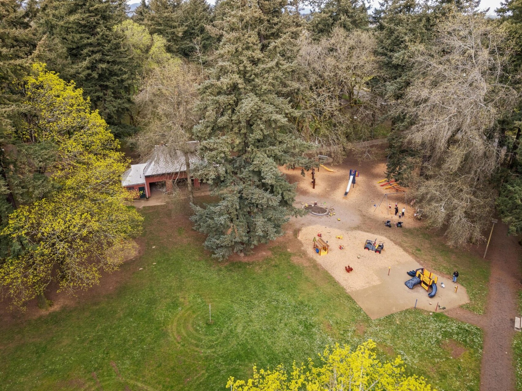 Aerial view of a park with a playground surrounded by tall trees, nestled in Portland's scenic landscape. The playground features slides, swings, and climbing structures. A wooden shelter is visible nearby, and several people are enjoying the area on a sunny day—prime for Portland Oregon real estate seekers.