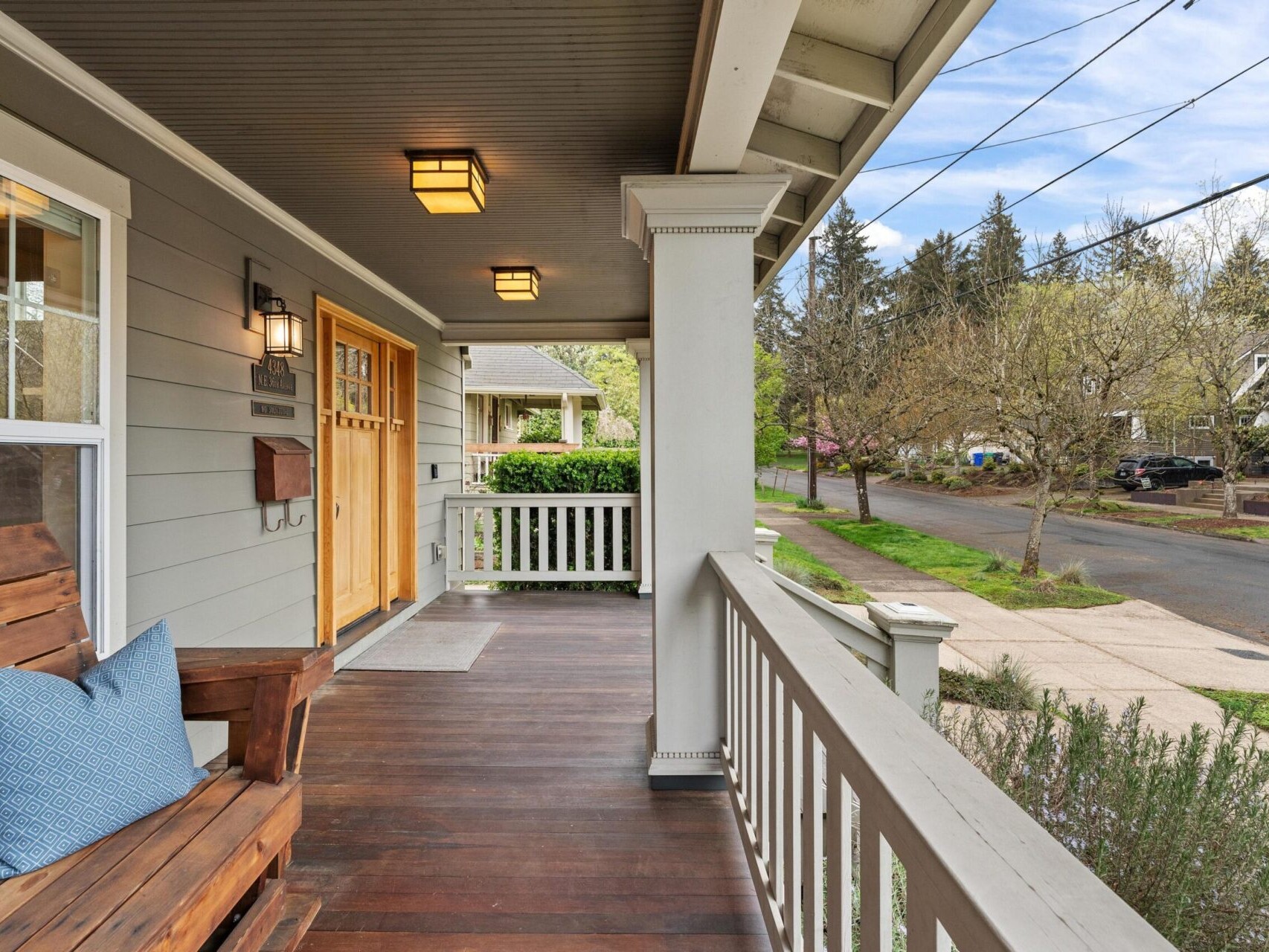 A cozy front porch in Portland, Oregon real estate features a wooden bench with a blue cushion, two wooden doors with windows, and views of a tree-lined street. The porch’s white railing and overhead lights create an inviting atmosphere cherished by any top real estate agent.