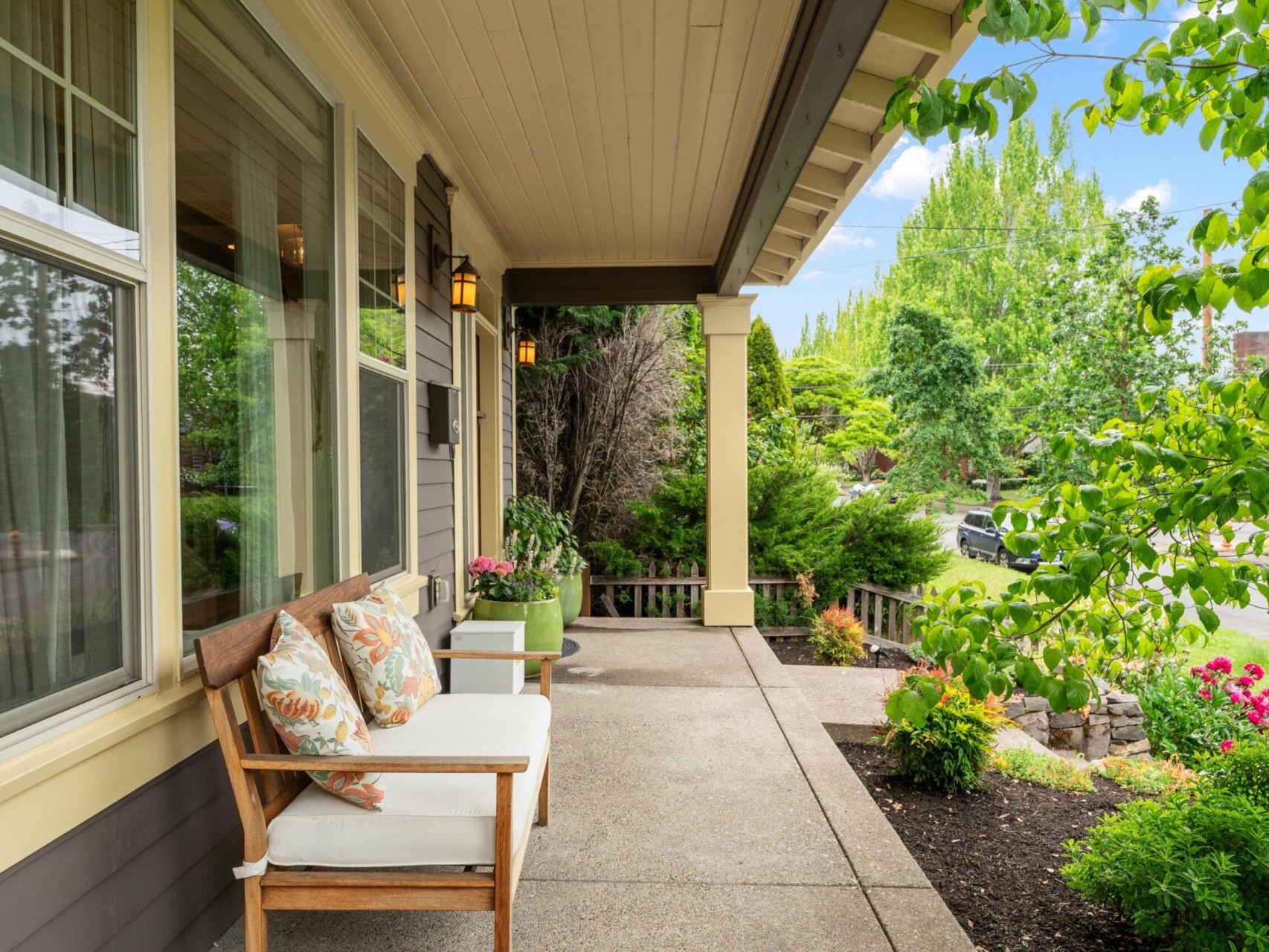 A cozy front porch with a wooden bench and two cushions, flanked by small green tables, overlooks a lush garden in a prime Portland Oregon real estate location. The clear blue sky enhances this inviting outdoor setting, perfect for anyone exploring options with a top real estate agent.