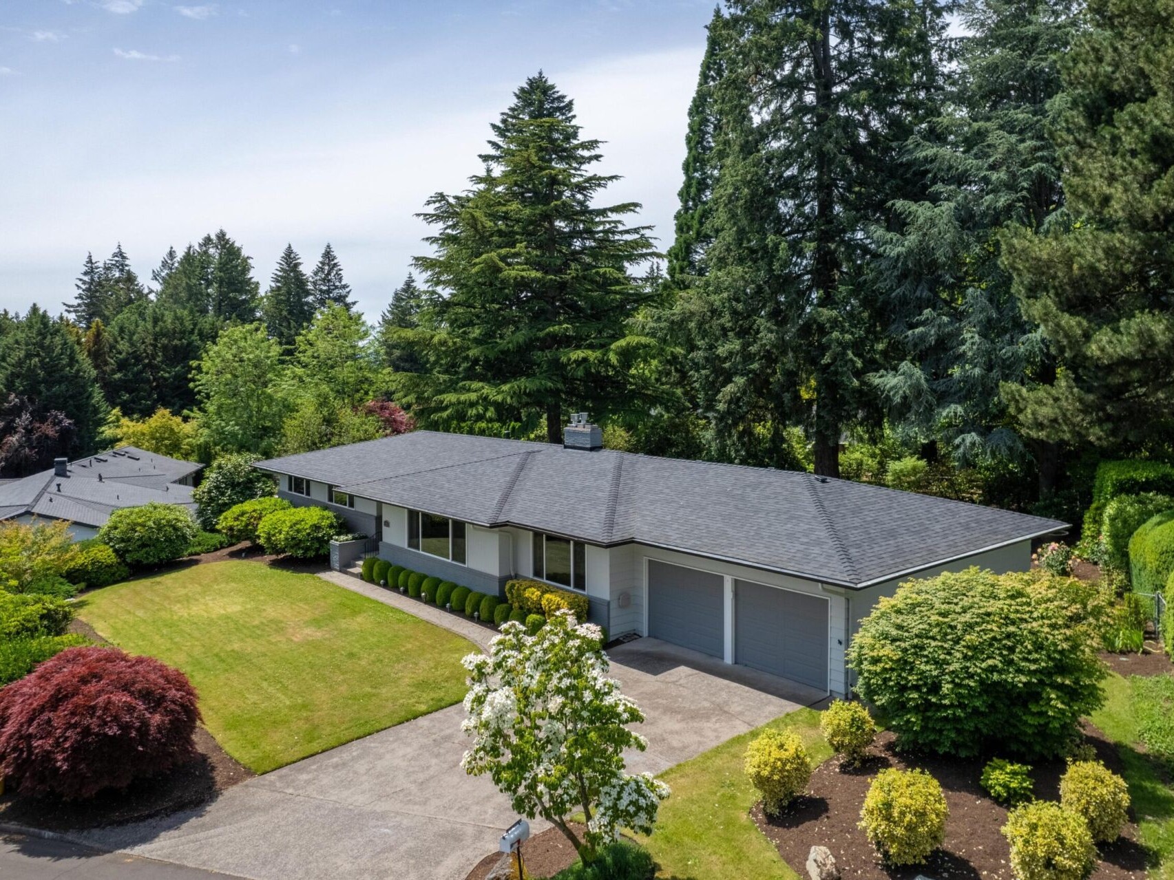 Aerial view of a single-story house with a dark roof, surrounded by lush greenery and manicured landscaping. The property features a driveway, garage, and various trees and shrubs, with a clear sky above.