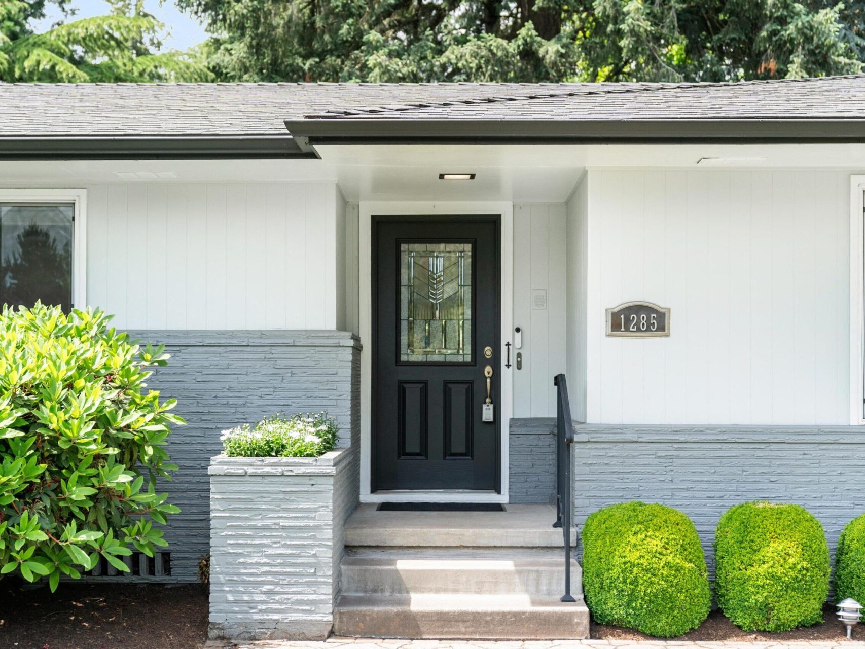 A modern house entrance with a black front door and white walls. The pathway is flanked by trimmed green bushes, and the house number 1265 is displayed near the door. Lush trees are visible in the background.