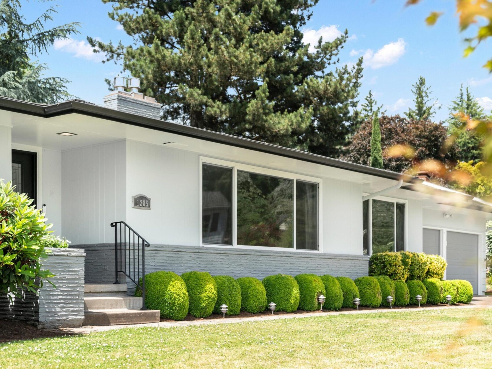 A modern single-story house with large windows, white and gray exterior, surrounded by neatly trimmed green bushes and lush trees. A concrete walkway leads to the entrance, and a garage is visible on the right. The sky is clear and blue.