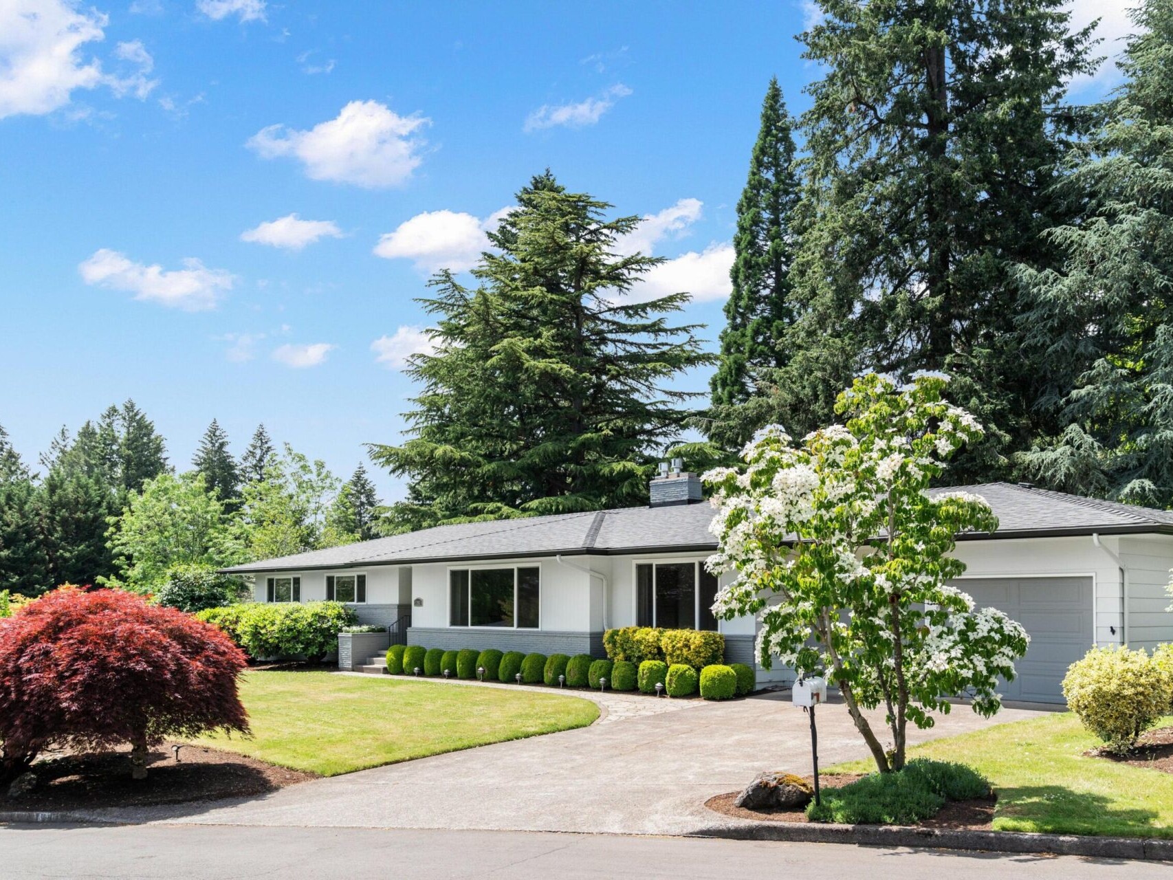 A single-story house with a gray roof and white exterior is surrounded by lush greenery and trees. A well-manicured lawn and driveway are in the foreground, with a clear blue sky overhead.