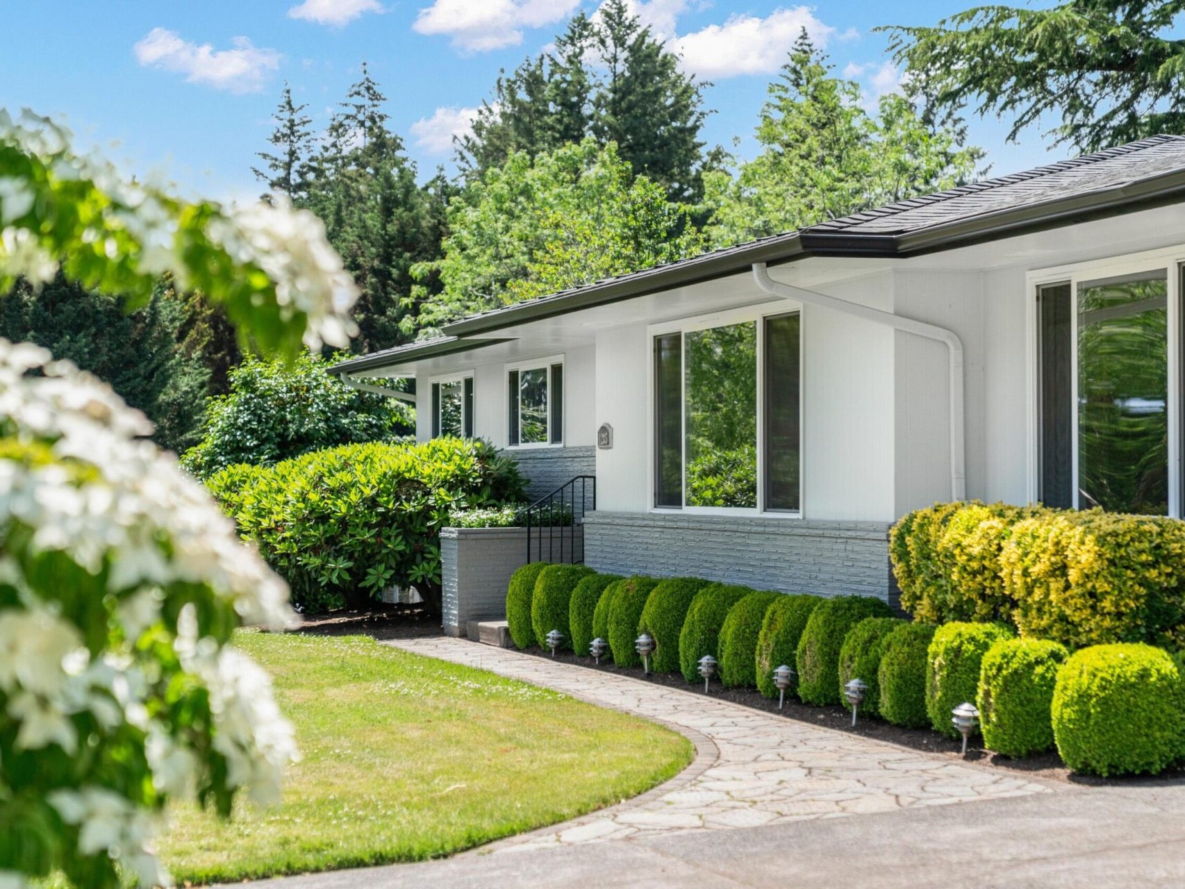 A well-maintained, single-story house with large windows, surrounded by green hedges and trees. A stone pathway curves through a manicured lawn, with white flowering plants in the foreground under a clear blue sky.