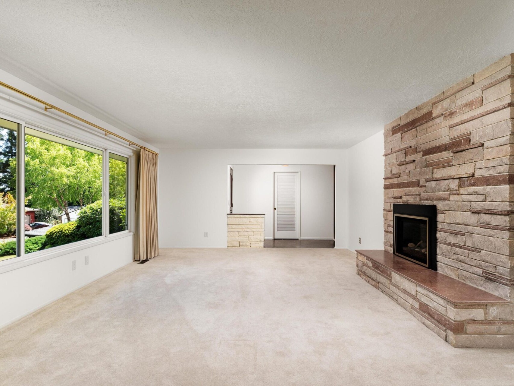 A spacious living room with beige carpet and a large window revealing a lush outdoor view. There’s a brick fireplace on the right wall and a doorway opposite the window. The room has neutral-toned walls and curtains.