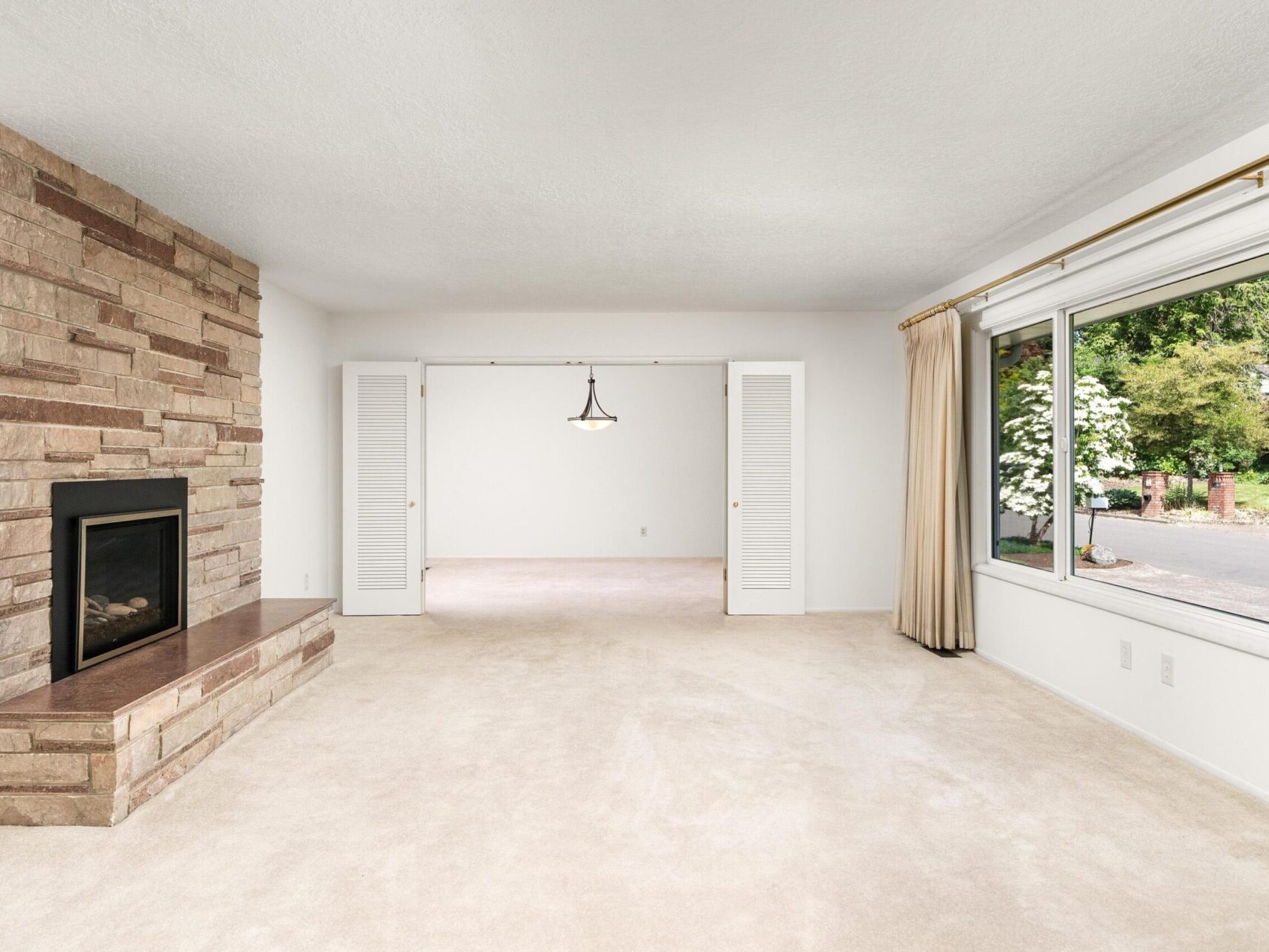 A spacious, empty living room with a large window showing greenery outside. A stone fireplace is on the left, and there are cream-colored walls and carpet. In the back, theres a chandelier and double doors leading to another room.