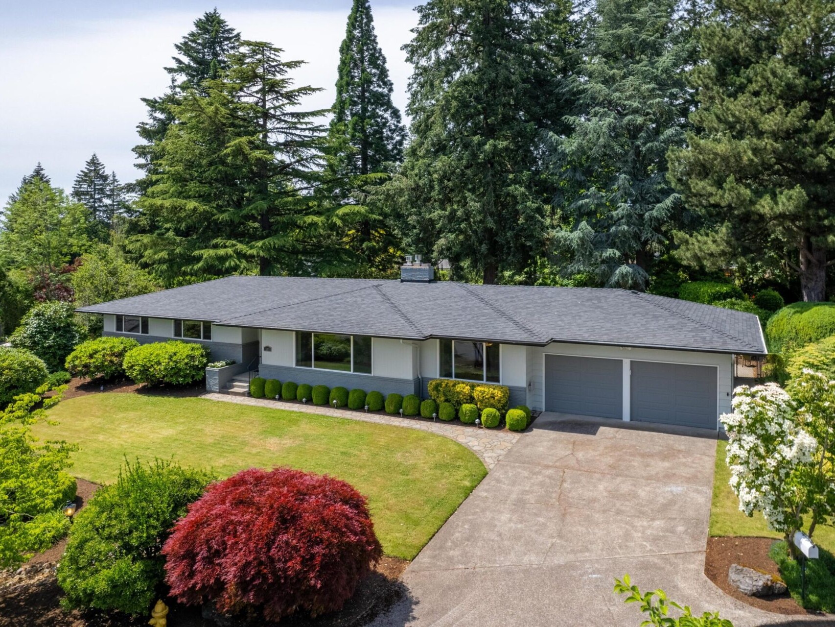 A single-story house with a gray roof and white exterior is surrounded by lush greenery and tall trees. A spacious driveway leads to a two-car garage, while well-maintained shrubs and a small lawn adorn the front yard.