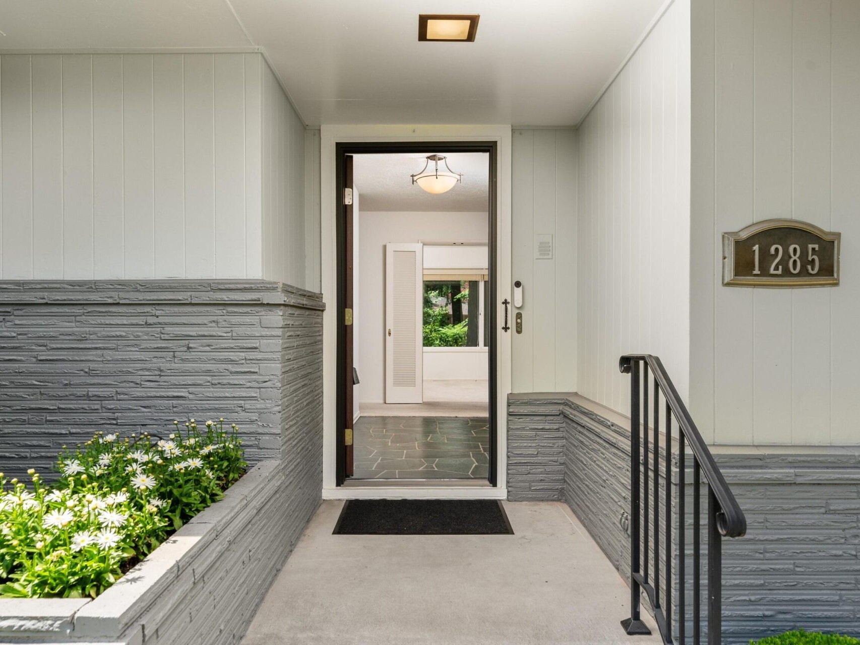 Front entrance of a modern Portland, Oregon real estate gem featuring a partially open door. The exterior has gray stone accents and the address number plate 1285. A small garden with white flowers is on the left, and a black mat welcomes you at the entrance, courtesy of your top real estate agent.