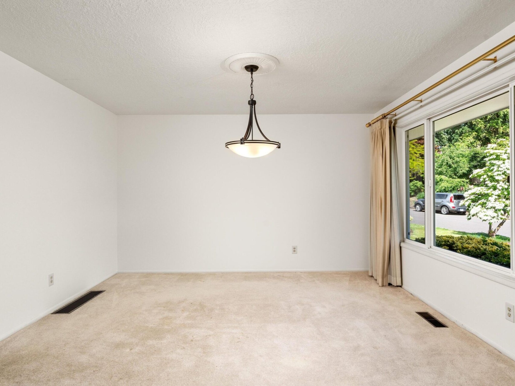 An empty room with beige carpet, white walls, a ceiling light fixture, and a large window with beige curtains. The window overlooks a street with parked cars and greenery.