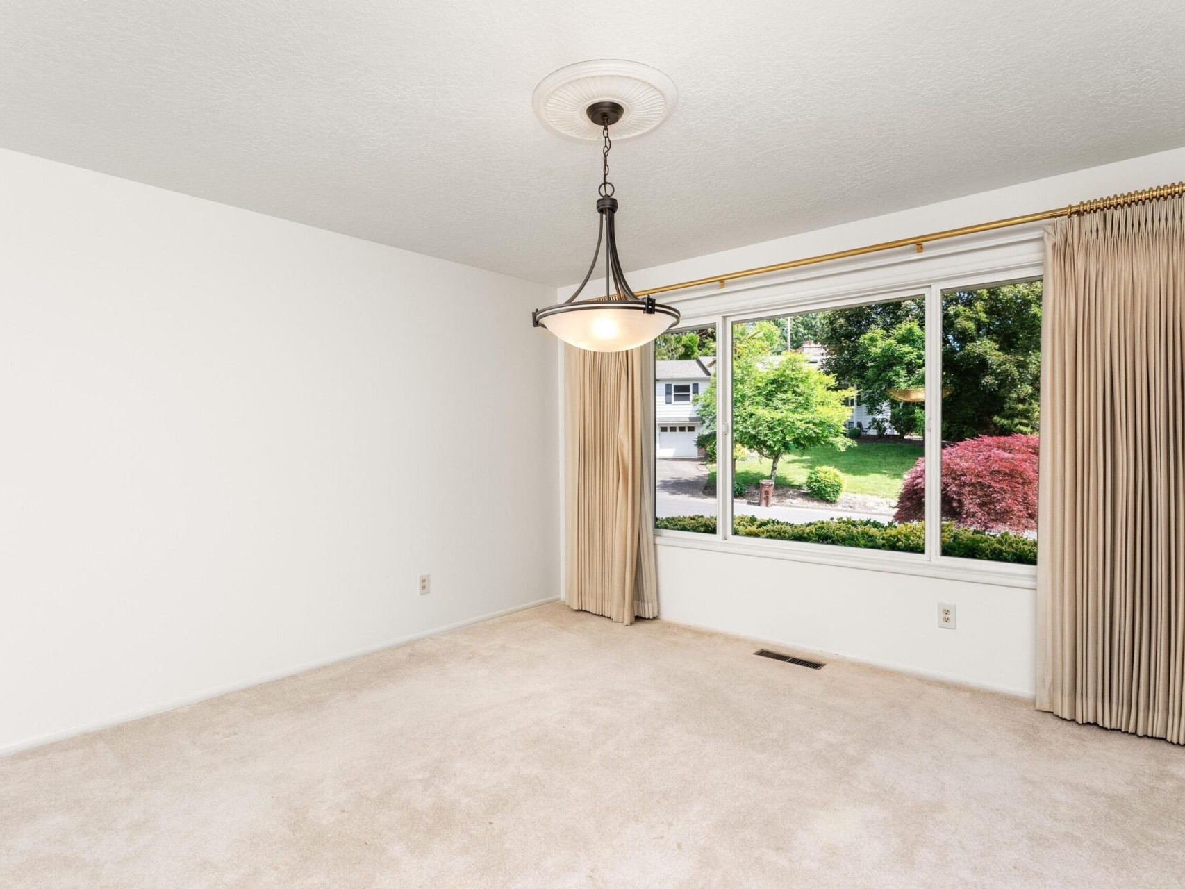 An empty room with white walls and beige carpet. A large window with closed beige curtains offers a view of a lush green garden. A modern ceiling light fixture hangs from the ceiling.