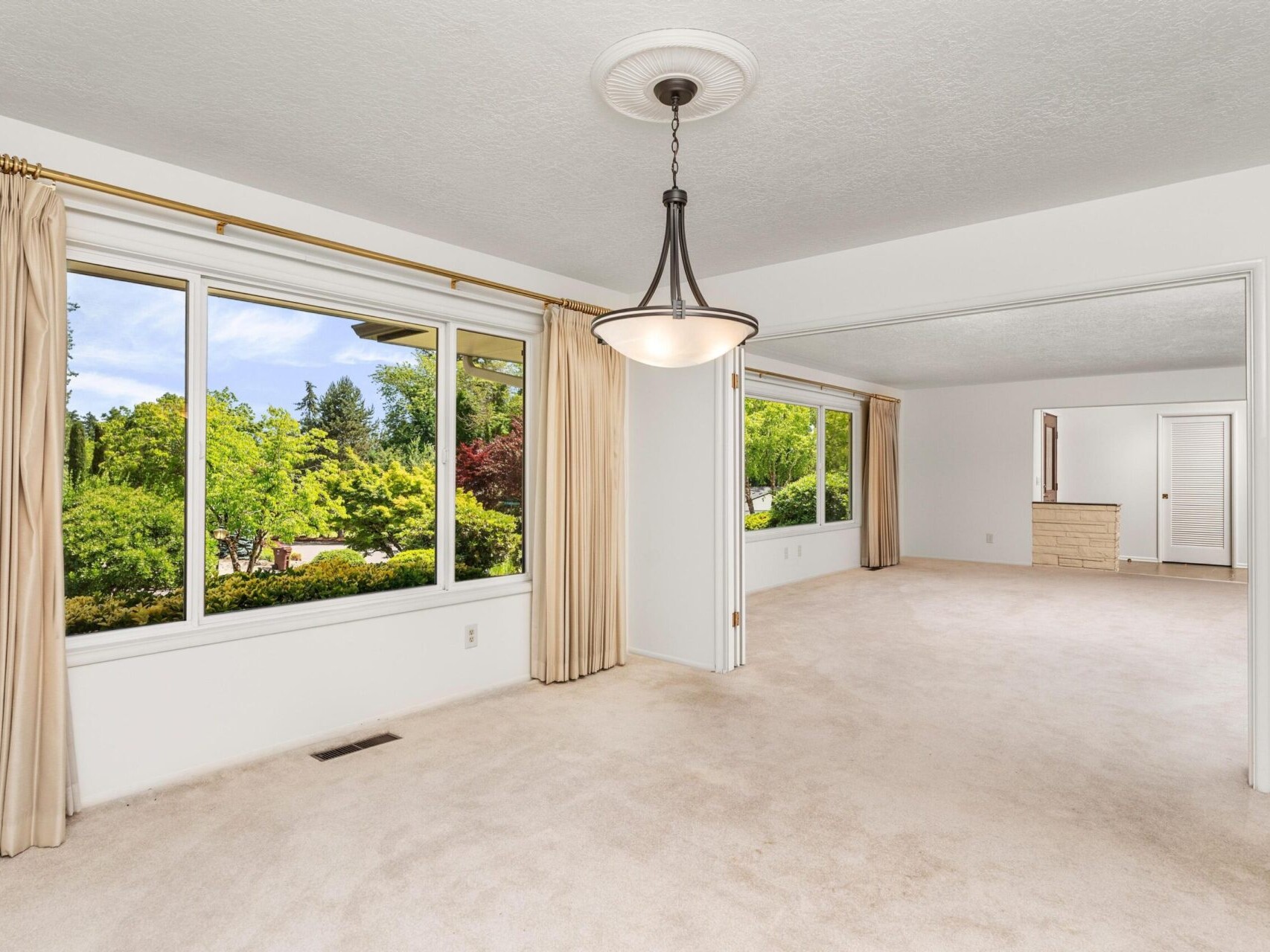 A bright, spacious living area with large windows showcasing green trees outside. The room features beige carpet, cream walls, and a modern hanging light fixture. Off to the side, a doorway leads to another room with a wooden dresser.