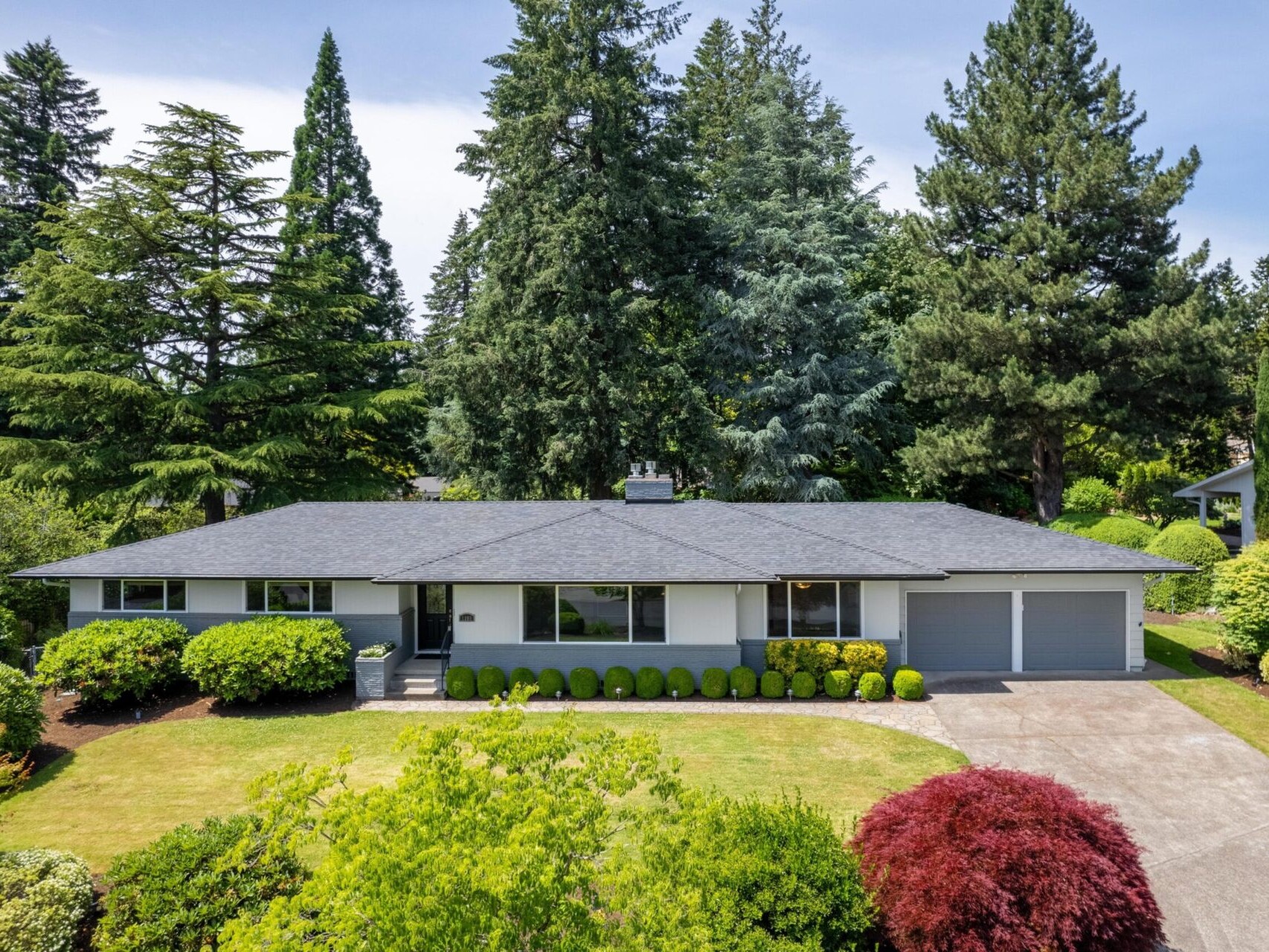 Aerial view of a single-story house with a dark roof, surrounded by lush greenery and tall trees. The front yard features a lawn and various shrubs, while a driveway leads to a two-car garage.