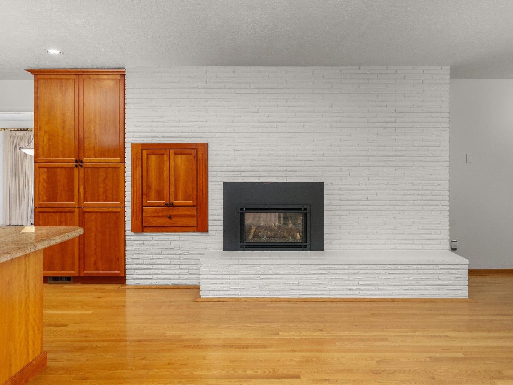 A modern living room with a white brick accent wall features a black fireplace. The floor is polished wood, and there are wooden cabinets to the left. A partially visible stone countertop is in the foreground on the left.