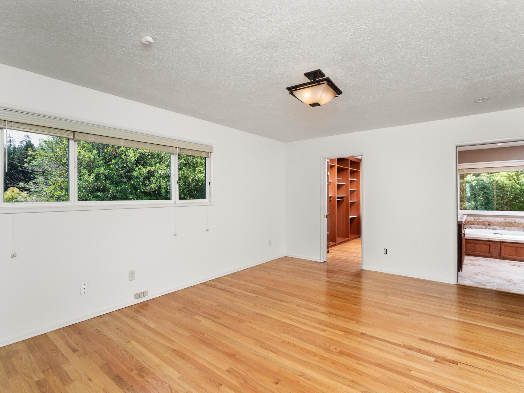 An empty room with hardwood floors, white walls, and a window displaying greenery outside. A doorway leads to another room with wooden shelves, and there is a light fixture on the ceiling.