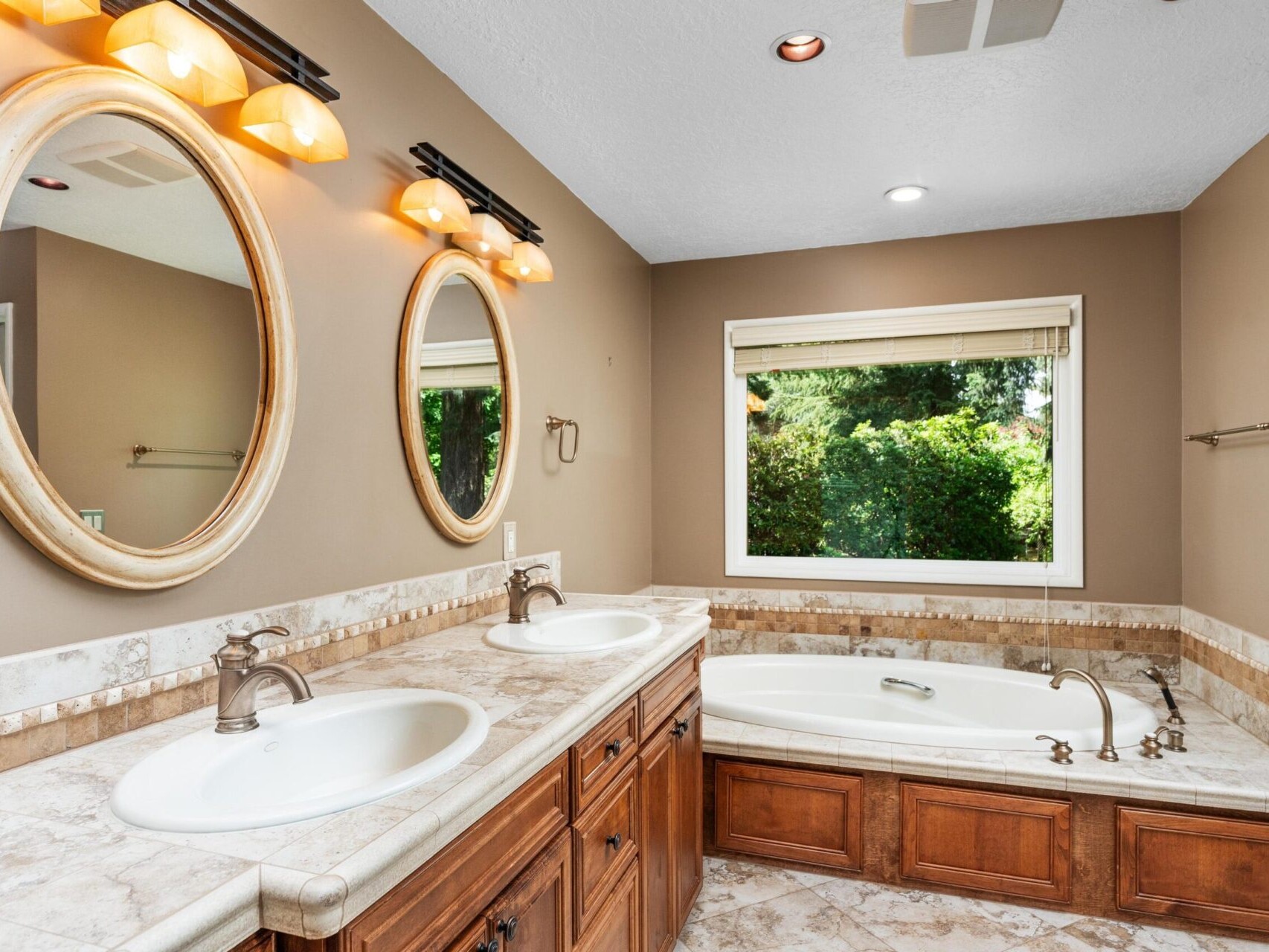 A bathroom with a dual sink vanity and two round mirrors. Marble countertops complement the wooden cabinets. A bathtub is set below a large window with garden views. Wall-mounted lights provide warm illumination.