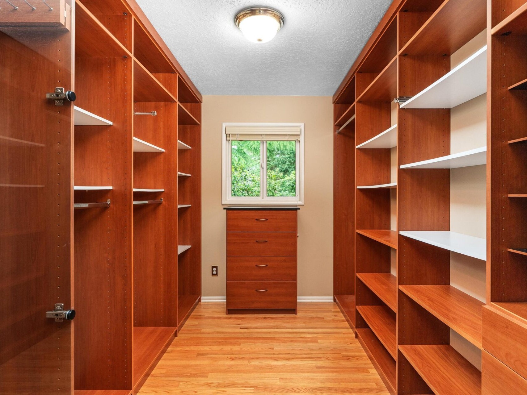 A wooden walk-in closet with open shelves and hanging rods on both sides, featuring a single chest of drawers beneath a small window at the end. The window shows a green, leafy view outside. The floor is made of light wood.