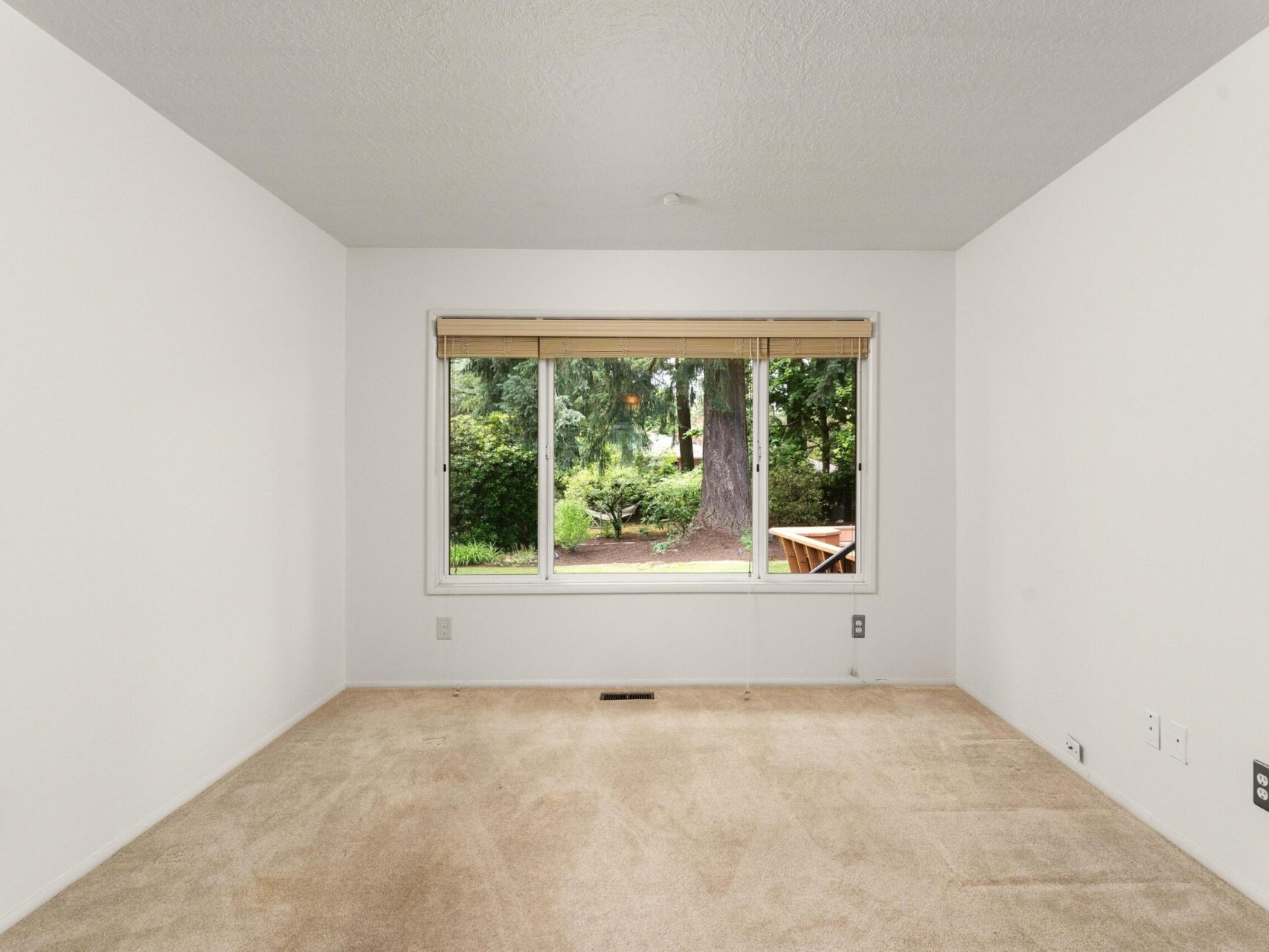 An empty room with light beige carpet, white walls, and a large window revealing a garden view. The window has a wooden frame and blind. A wooden bench is visible outside.