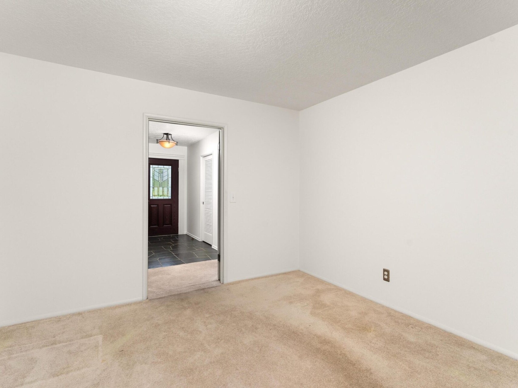 Minimalist room with off-white walls and beige carpet. An open doorway leads to a hallway with dark tile flooring and a visible wooden door in the distance. A single ceiling light fixture is in view.