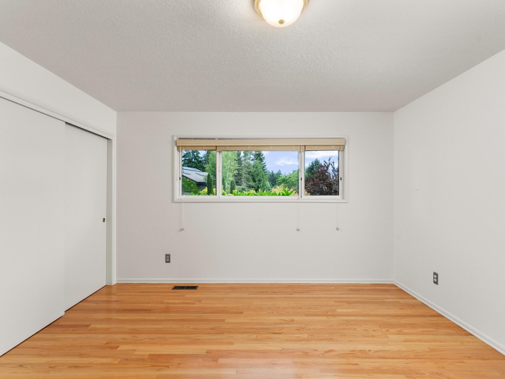 An empty room with wooden flooring, white walls, a ceiling light, and a small window with a view of trees and a cloudy sky. A sliding closet door is on the left wall.