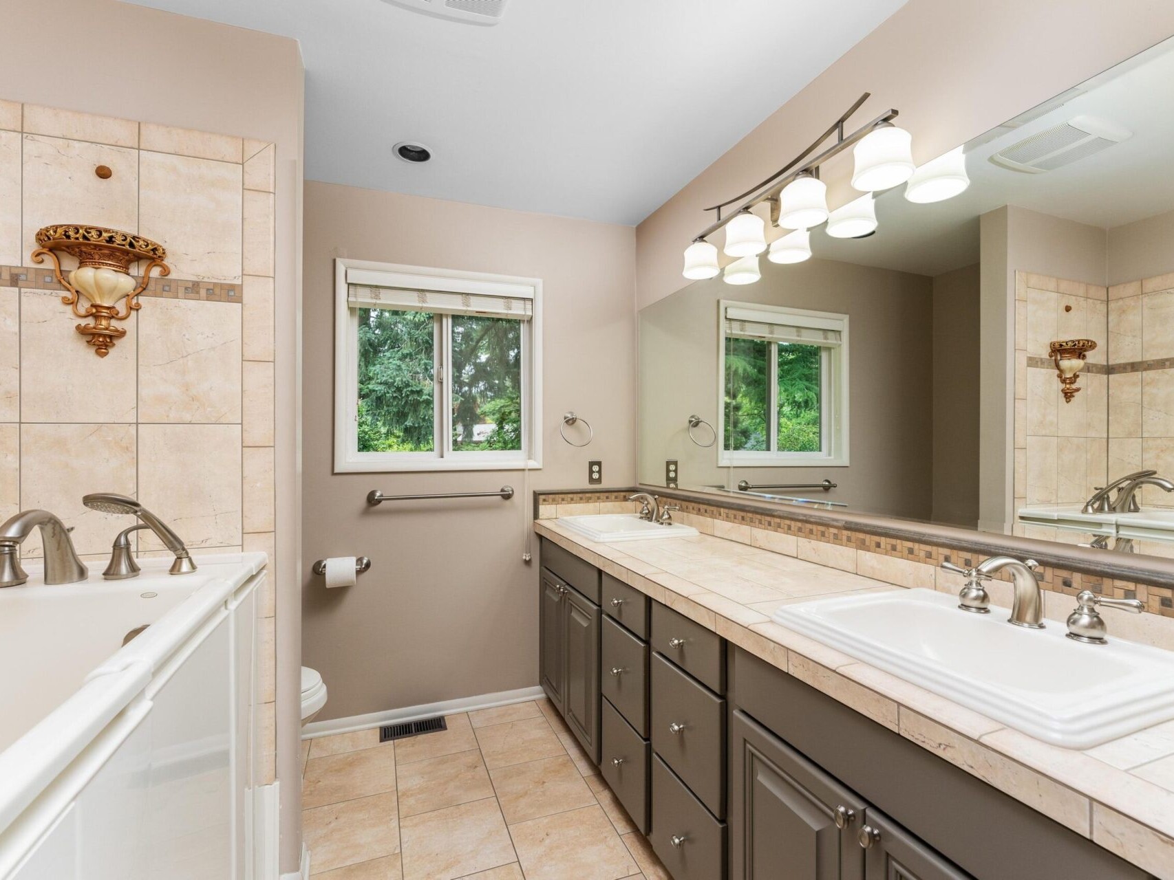A modern bathroom with a double sink vanity, large mirror, and wall-mounted light fixtures. The room features beige tiles, a window with a view of greenery, and a white bathtub next to the vanity.