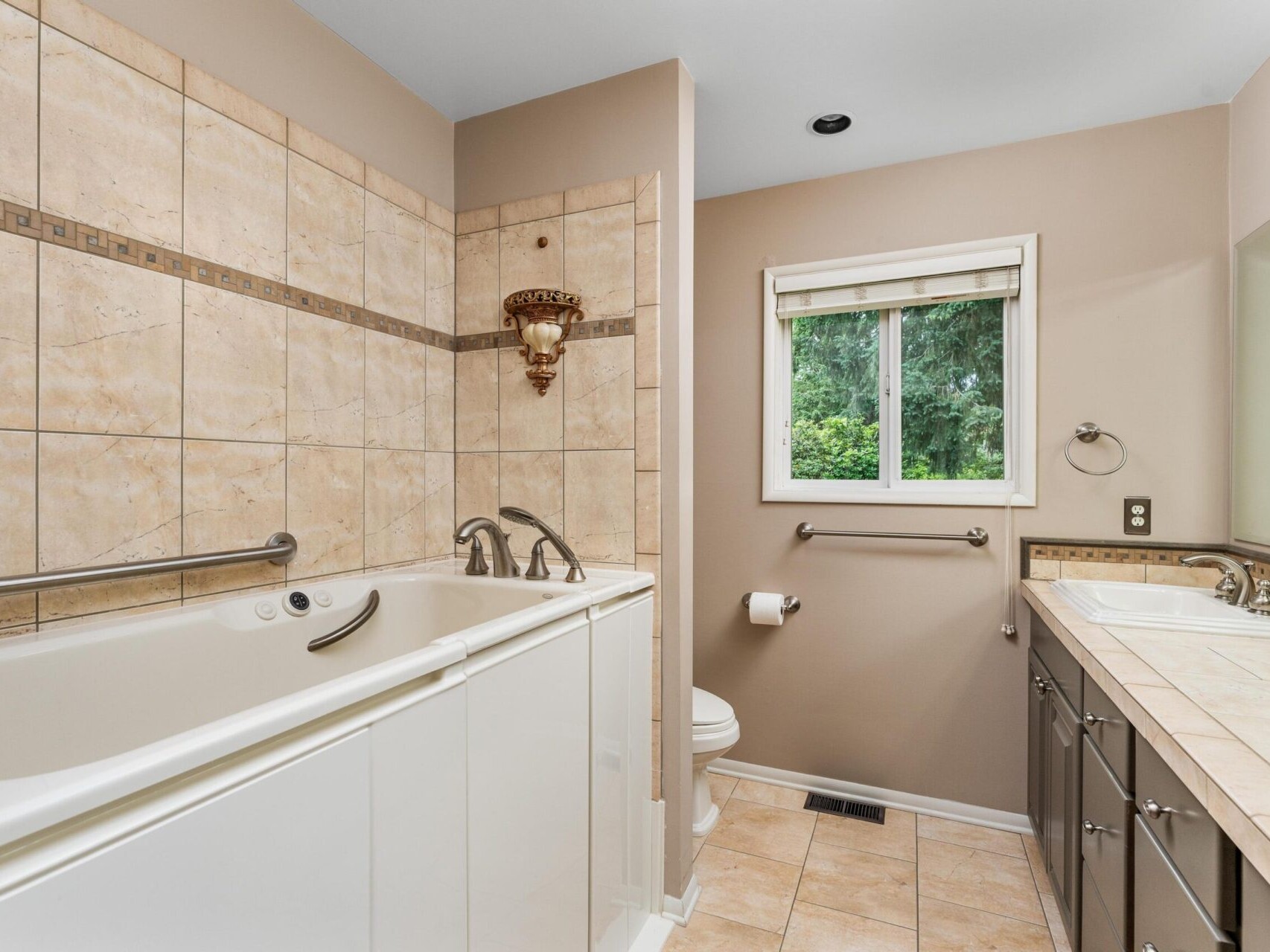 A bathroom with beige tile walls and floor features a walk-in bathtub with grab bars, a toilet, and a double sink vanity. A window offers a view of greenery outside, and a decorative light fixture is above the tub.