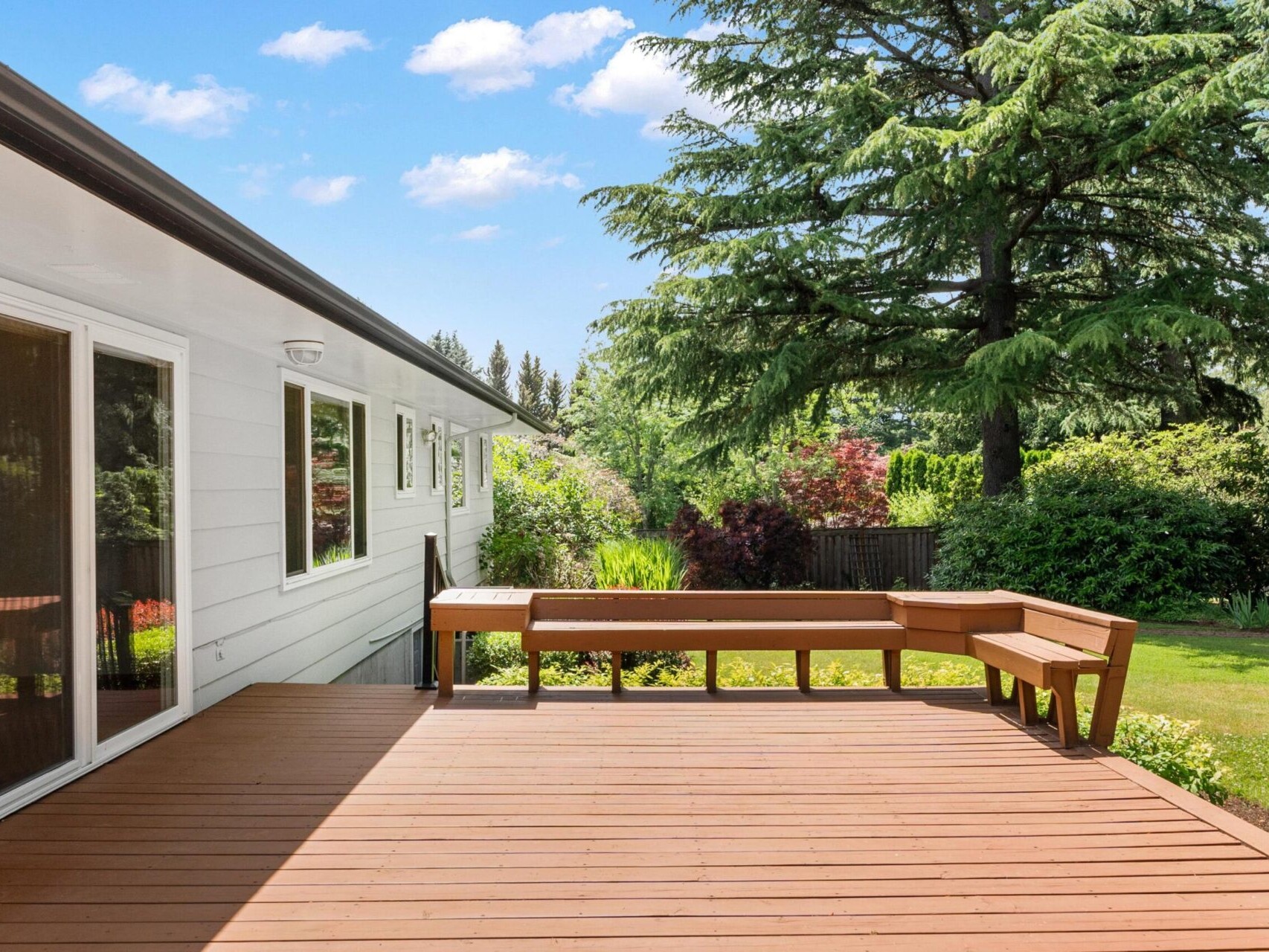 A wooden deck with attached seating overlooks a lush green backyard with various trees and shrubs. The side of a white house is visible on the left. Blue sky and fluffy clouds are in the background.