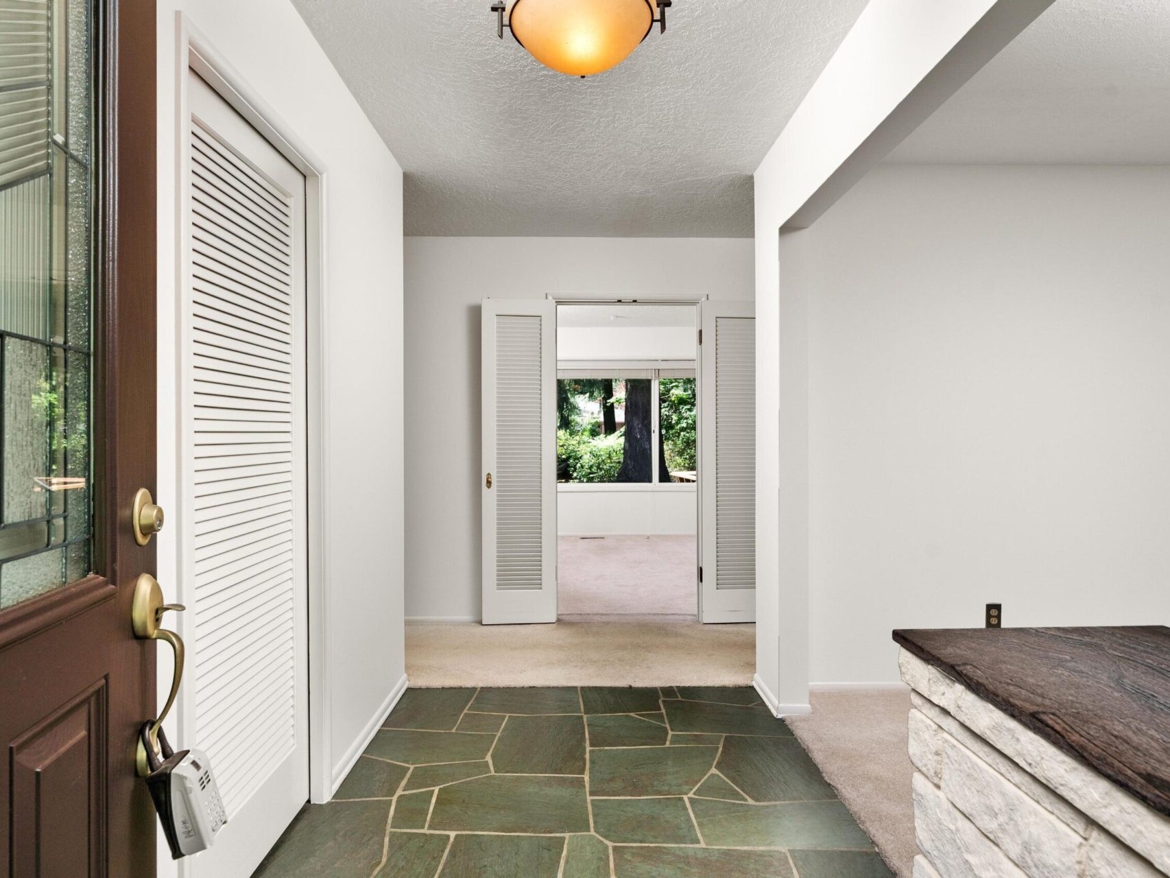 A bright, open hallway with white walls and a slate tile floor. A brown wooden door with glass panels is partially open on the left. Beyond, a room with carpet and a large window looking out to greenery. An amber light fixture hangs overhead.