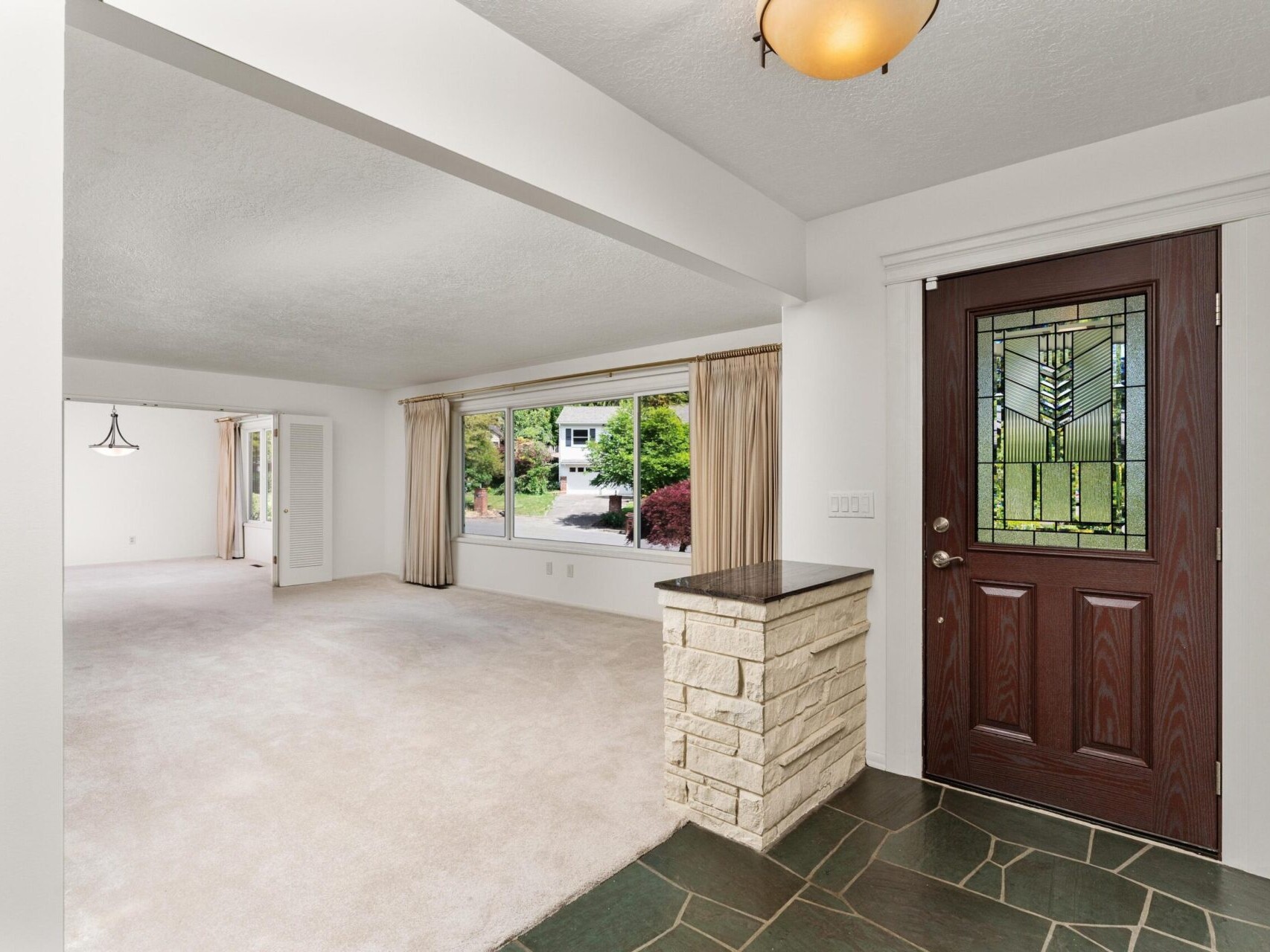 Spacious interior of a home with a tiled entryway, wooden front door featuring decorative glass, and a large living area. The room has carpeted floors, a wide window with curtains, and a glimpse of greenery outside.