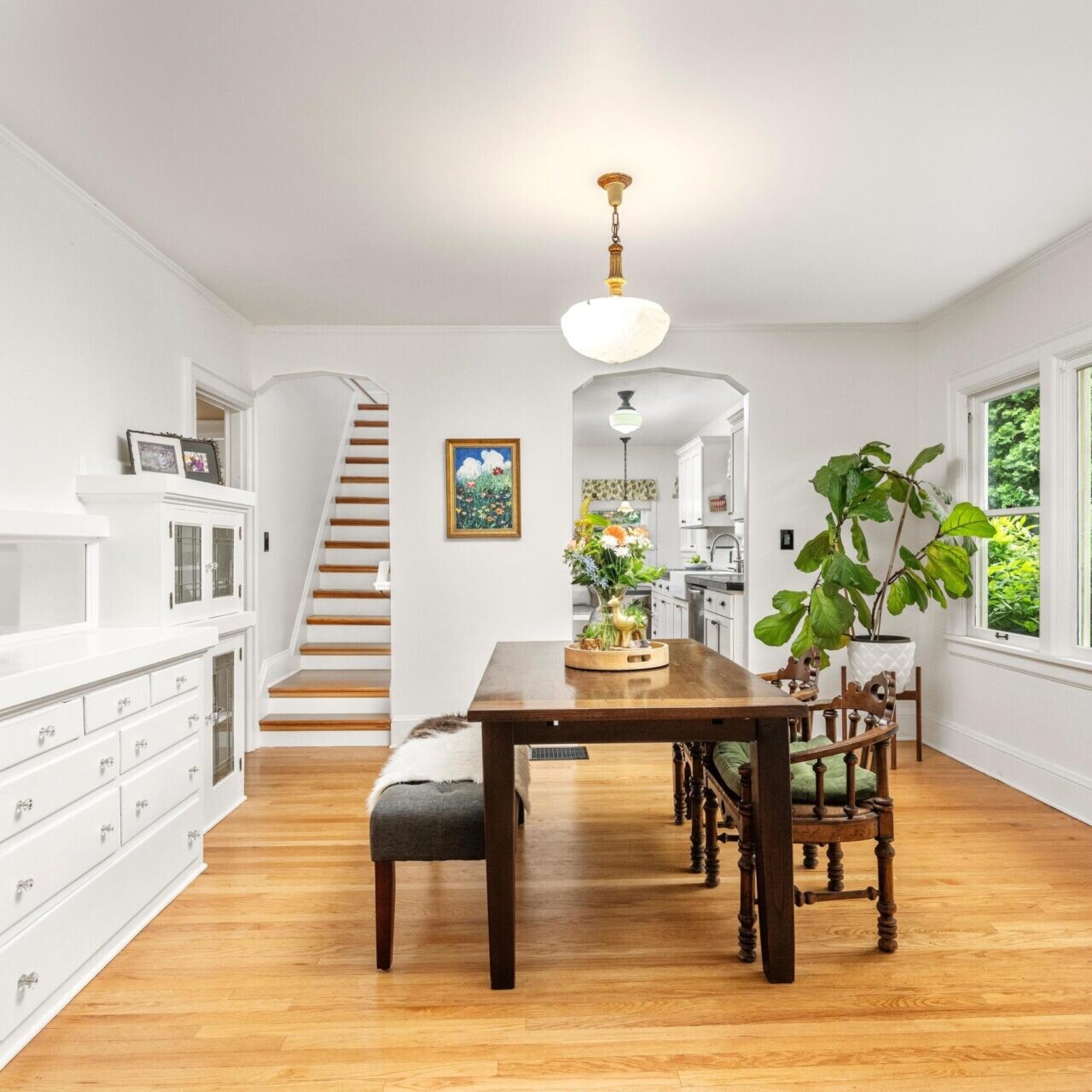 A bright dining room in a charming Portland home features a wooden table and chairs, expansive window views of lush greenery, and crisp white walls. To the left, you'll find a staircase, a plant by the window, and a cabinet with glass doors. Nearby hangs an art piece close to the stairs.