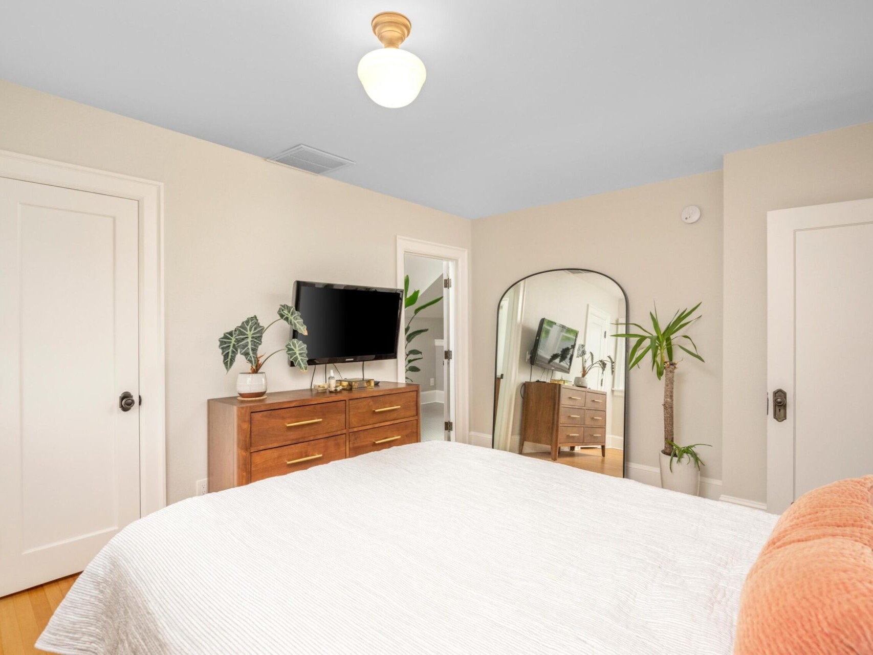 A bright bedroom in a Portland Oregon real estate gem features a large bed with white bedding and a peach cushion. A wooden dresser with a TV adorns the wall, complemented by a tall mirror and potted plants. The room's white walls, wooden floors, and ceiling light showcase its elegant charm.