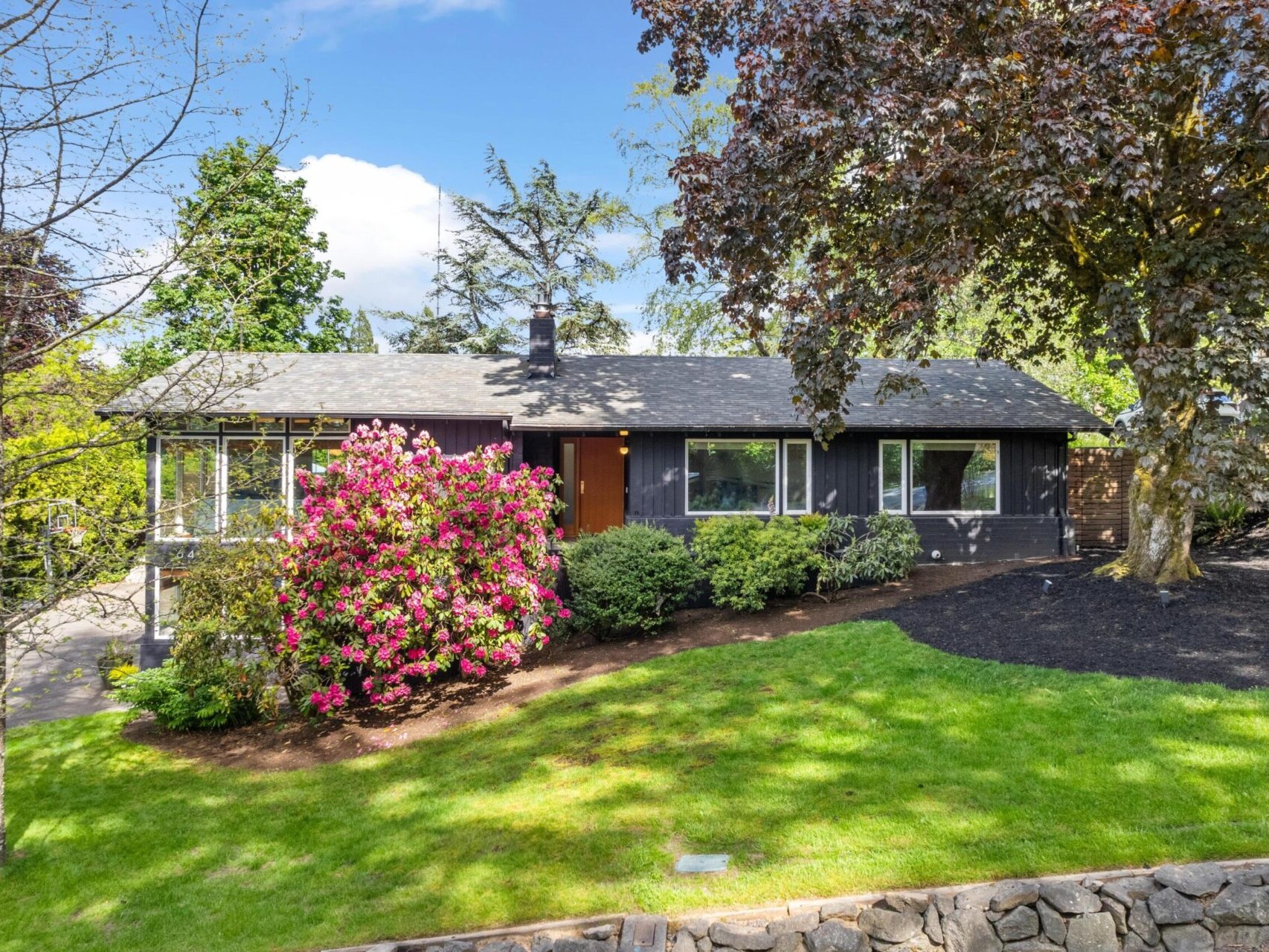 A charming single-story house with a dark exterior, surrounded by lush greenery. A vibrant pink flowering bush is near the main entrance, and a stone boundary wall lines the grassy front yard under a bright blue sky.