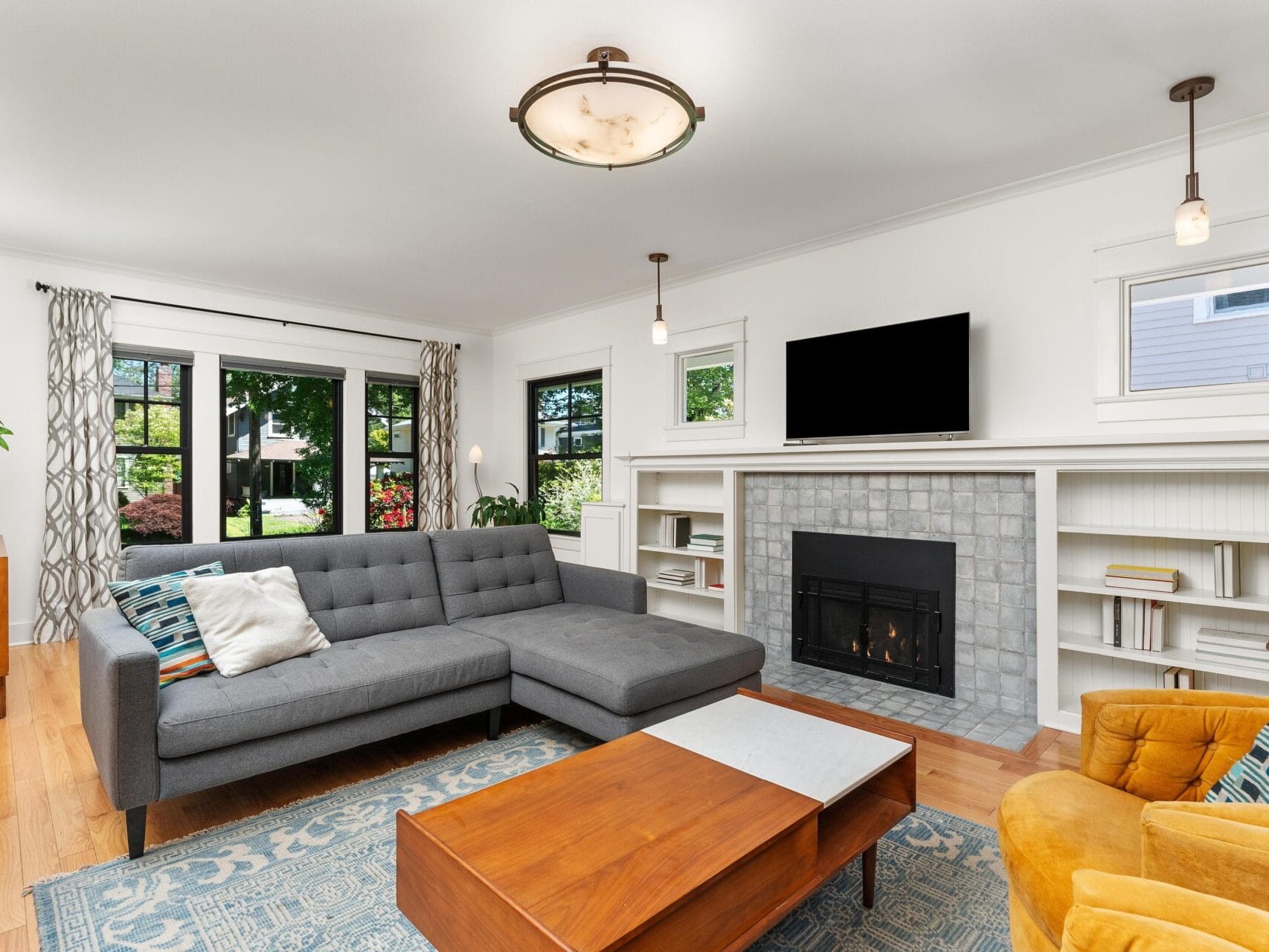 A modern living room with a gray sectional sofa, a wooden coffee table, and a fireplace with a flat-screen TV above it. Bookshelves flank the fireplace. A patterned rug and an orange armchair add color. Large windows let in natural light.