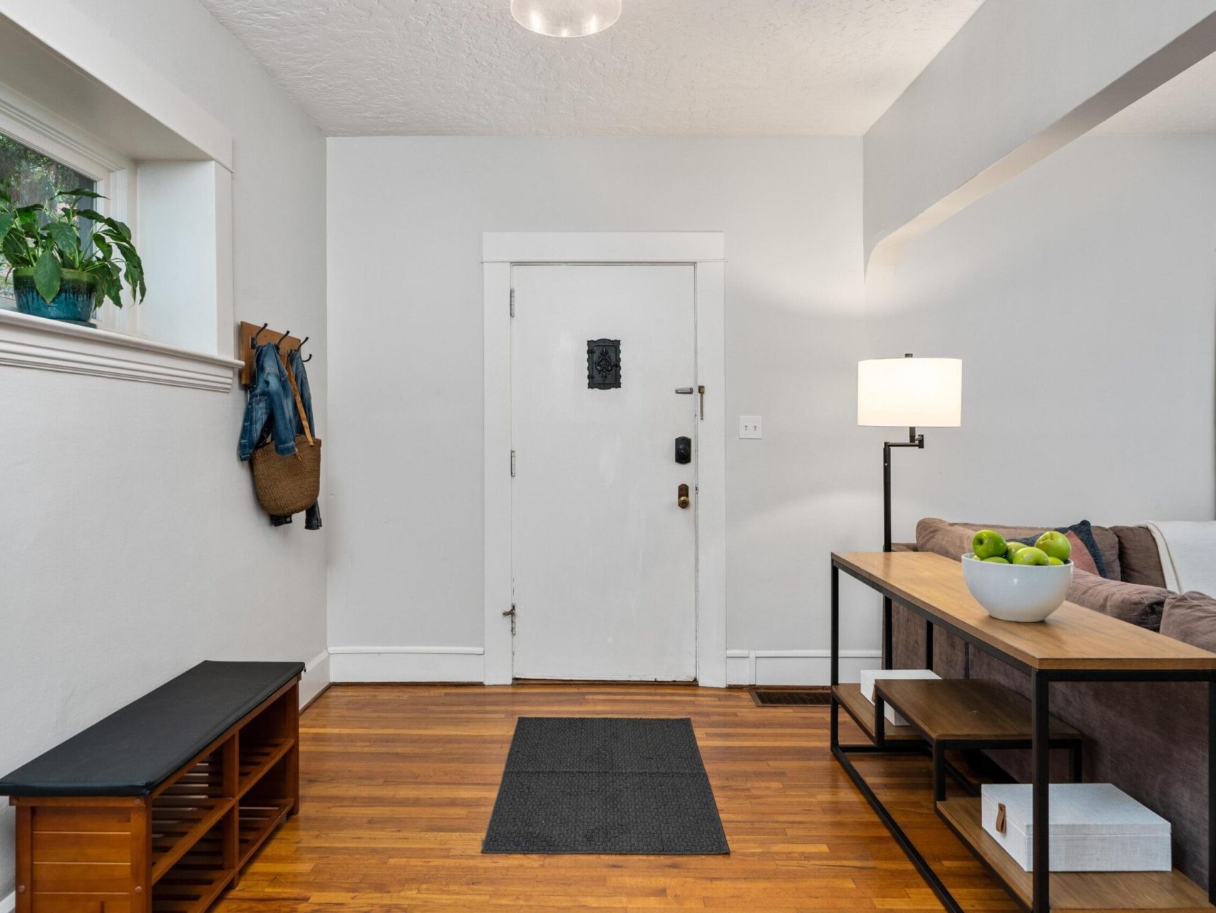 A bright living room with a white front door, a black bench, a small coat rack with jackets hanging on the wall, and a table with a lamp, a bowl of green apples, and shelf storage. The walls are light-colored, and the floor is wooden.