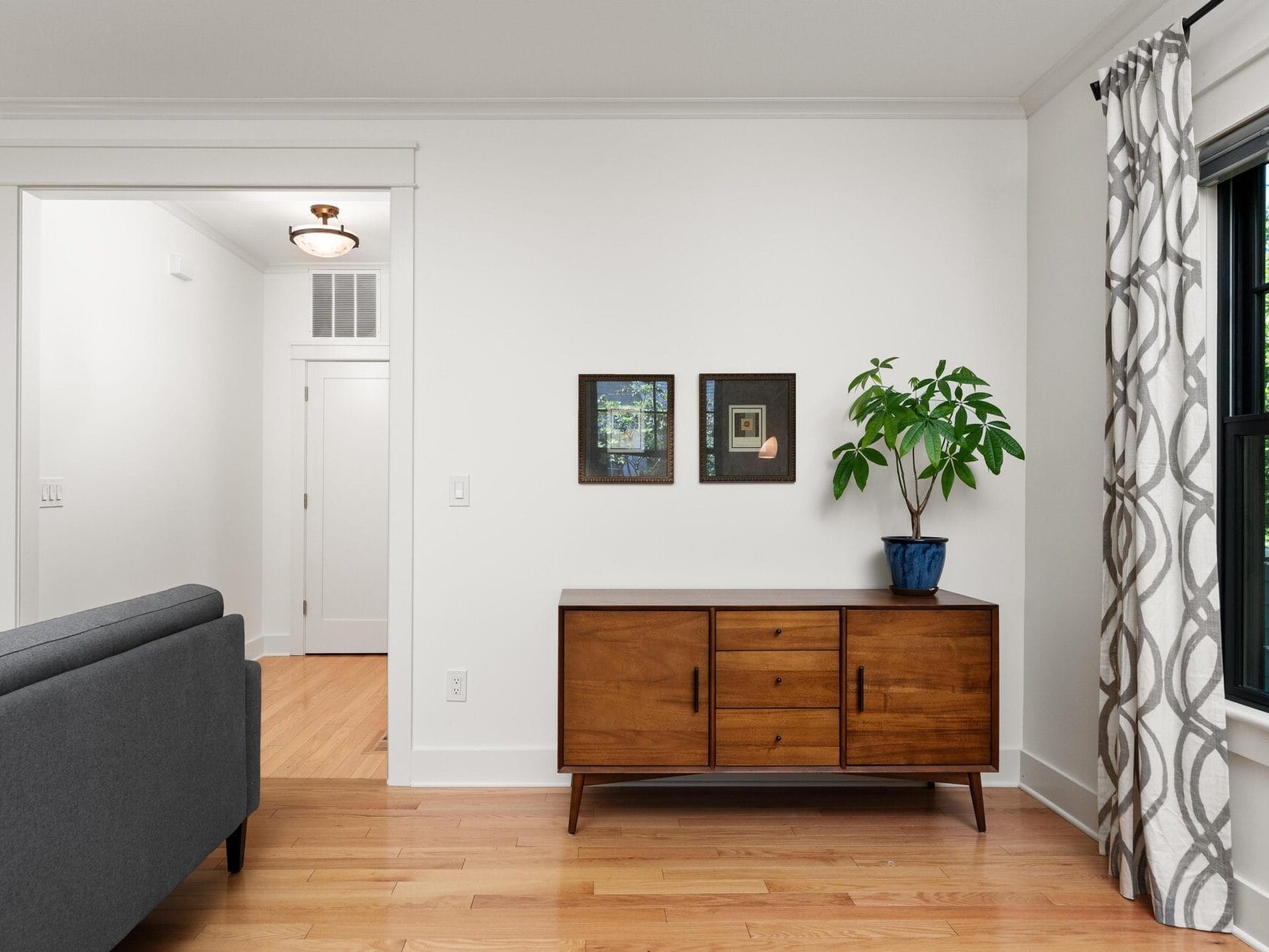 A minimalist living room with a mid-century modern wooden sideboard, decorated with small framed artwork and a potted plant. A gray sofa and patterned curtains are also visible, with a doorway in the background. Bright natural light illuminates the space.
