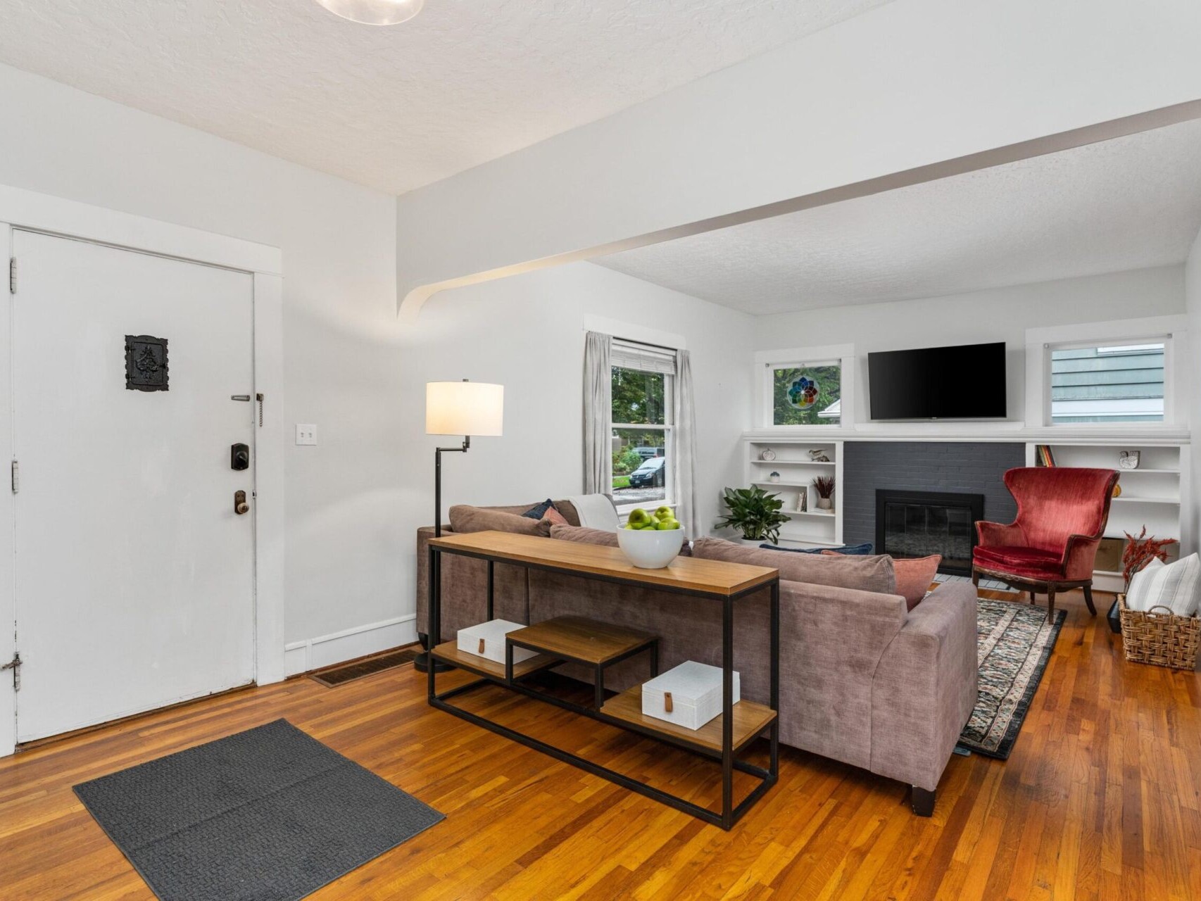 A cozy living room with hardwood floors featuring a gray sofa, a modern wooden coffee table, and a red armchair. Theres a fireplace with a TV above it, shelves on either side, and windows providing natural light. A potted plant and a floor lamp adorn the space.