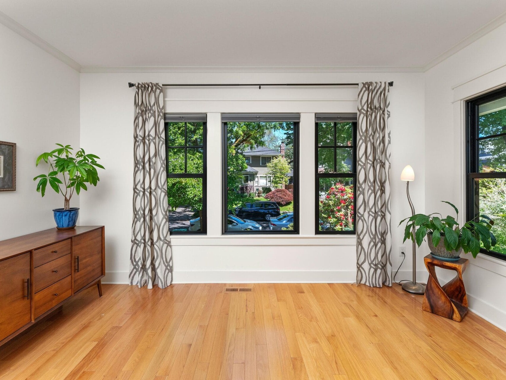 A bright room with a large window showcasing a garden view. The room features light wood flooring, a wooden sideboard with a potted plant, patterned curtains, and another plant near a floor lamp in the corner.