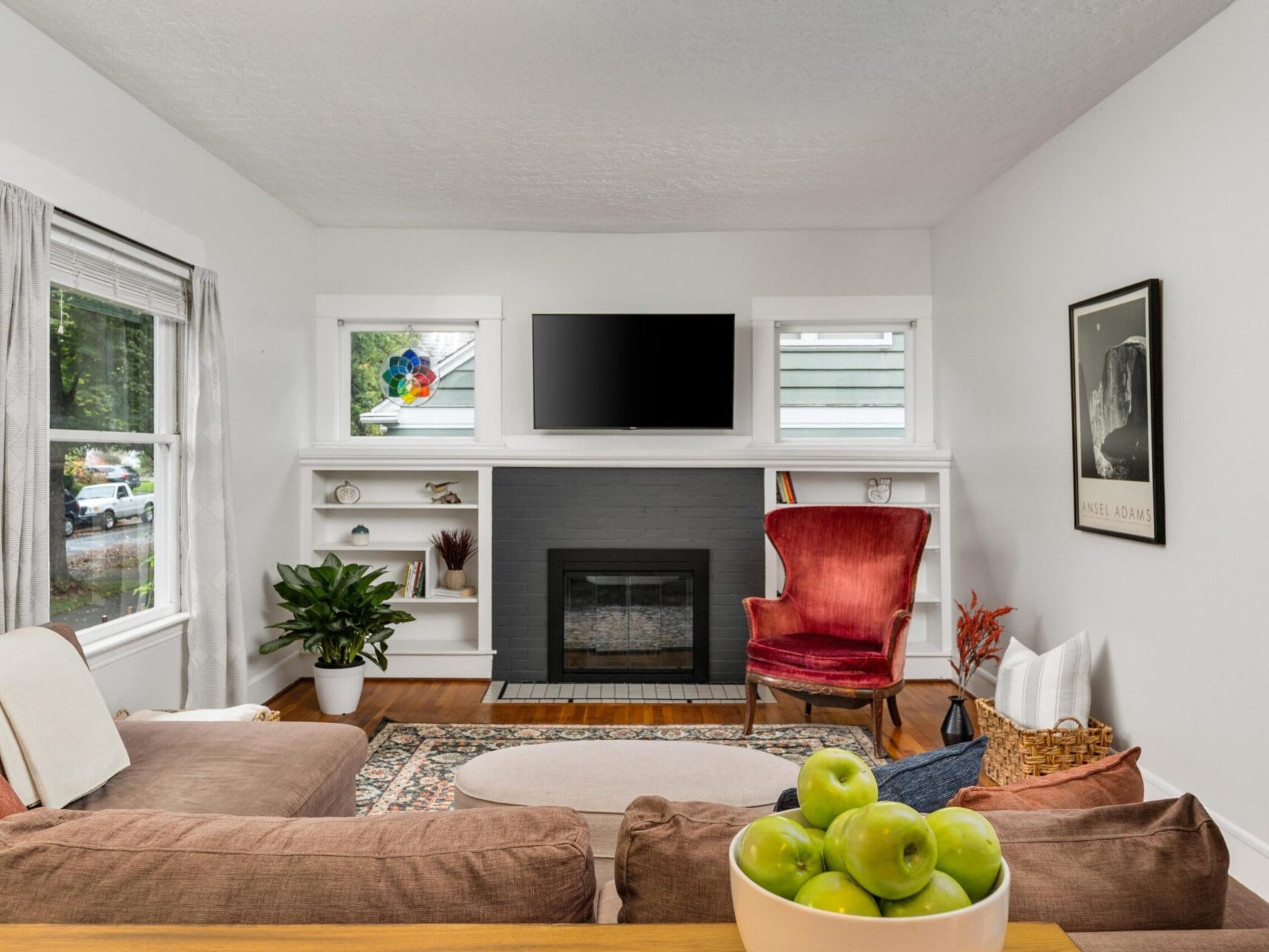 A cozy living room with a fireplace, TV, and built-in shelves. A red velvet chair faces a brown sectional sofa adorned with cushions. A basket of apples sits on the table. A houseplant and wall art add charm to the space. Natural light streams in.